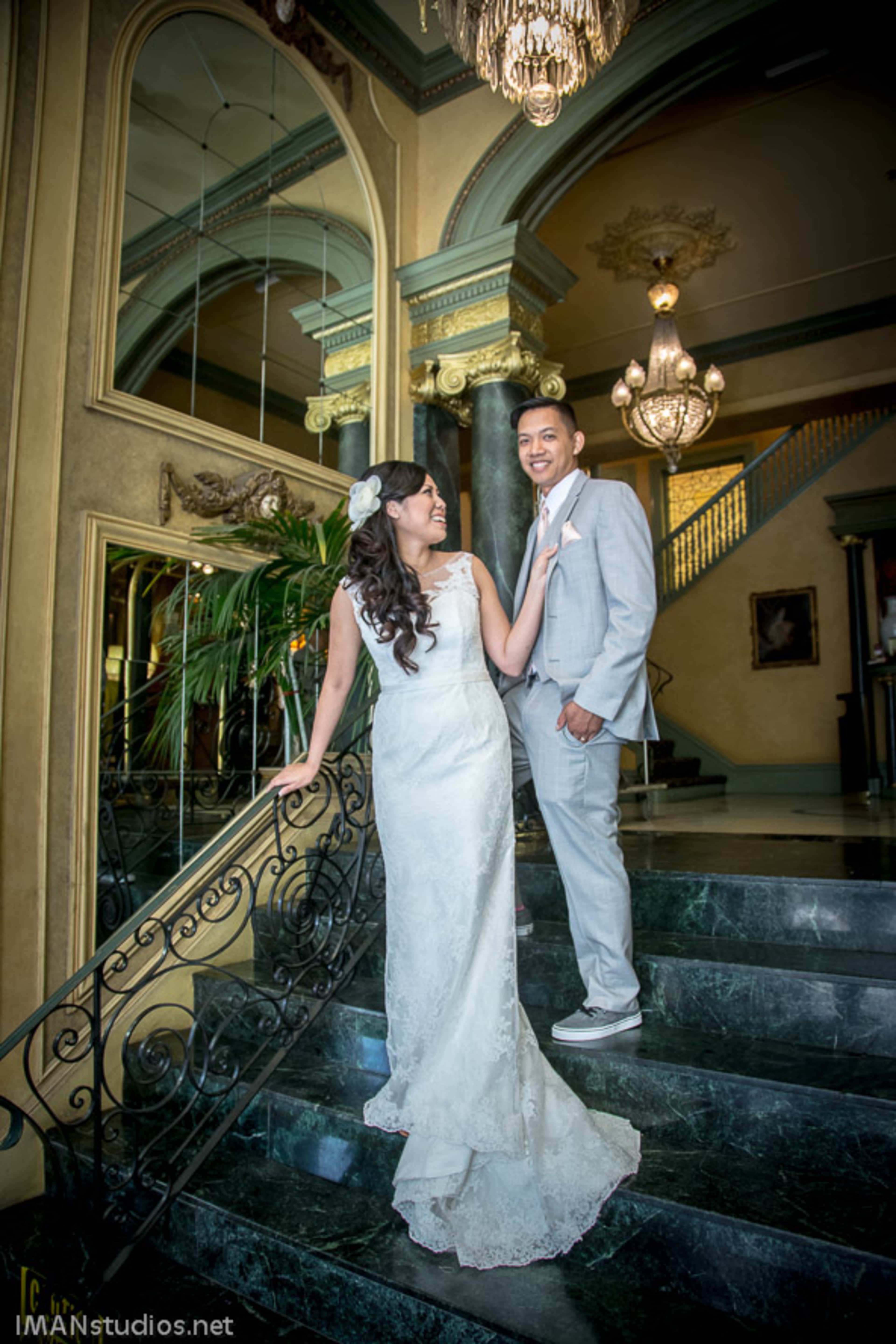 A bride in a white dress poses on a staircase with a groom in a light gray suit and sneakers, surrounded by elegant architectural elements.