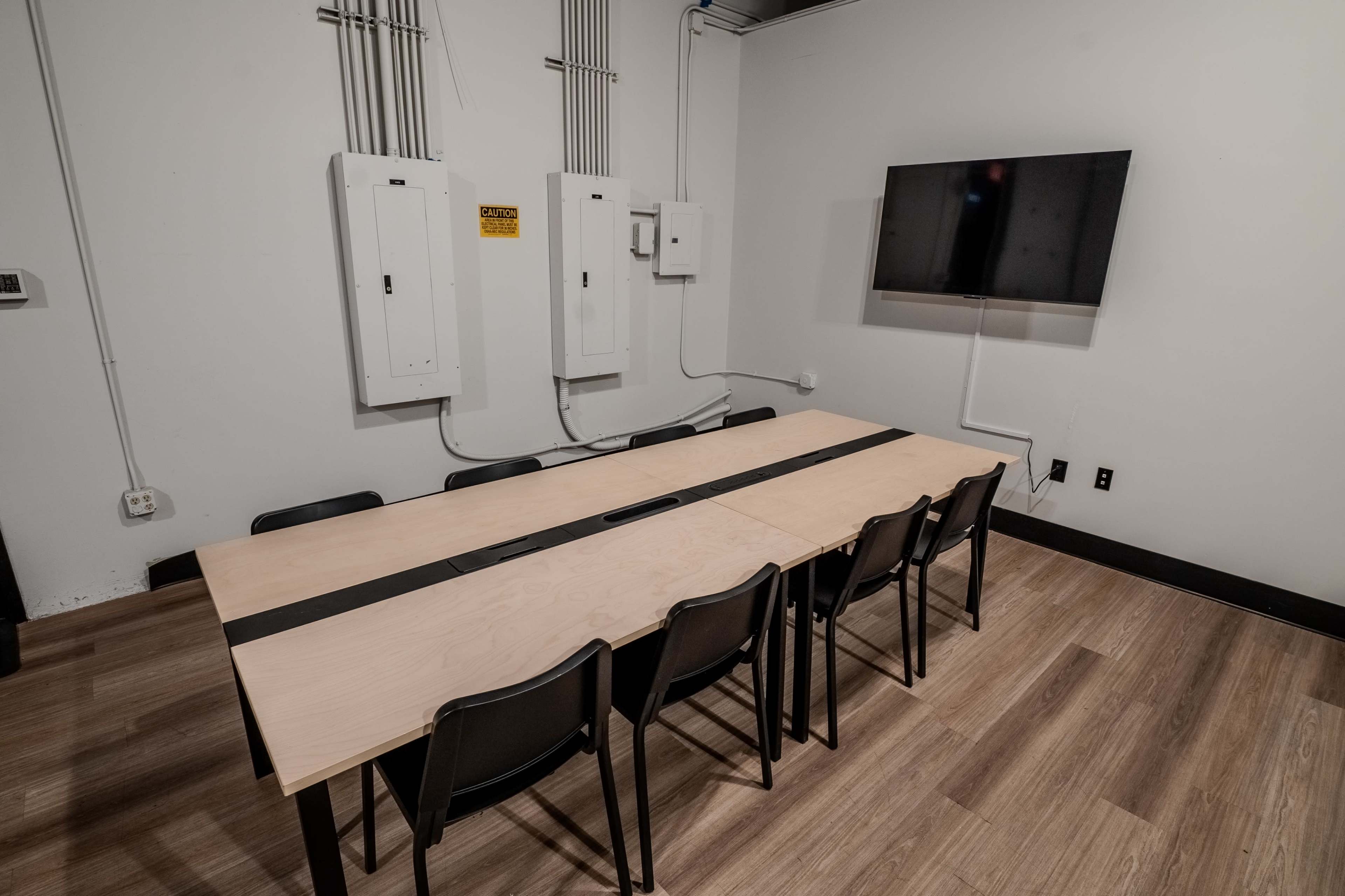 The image shows a conference room featuring a long wooden table with six black chairs on either side, a wall-mounted television, and electrical panels in the background.