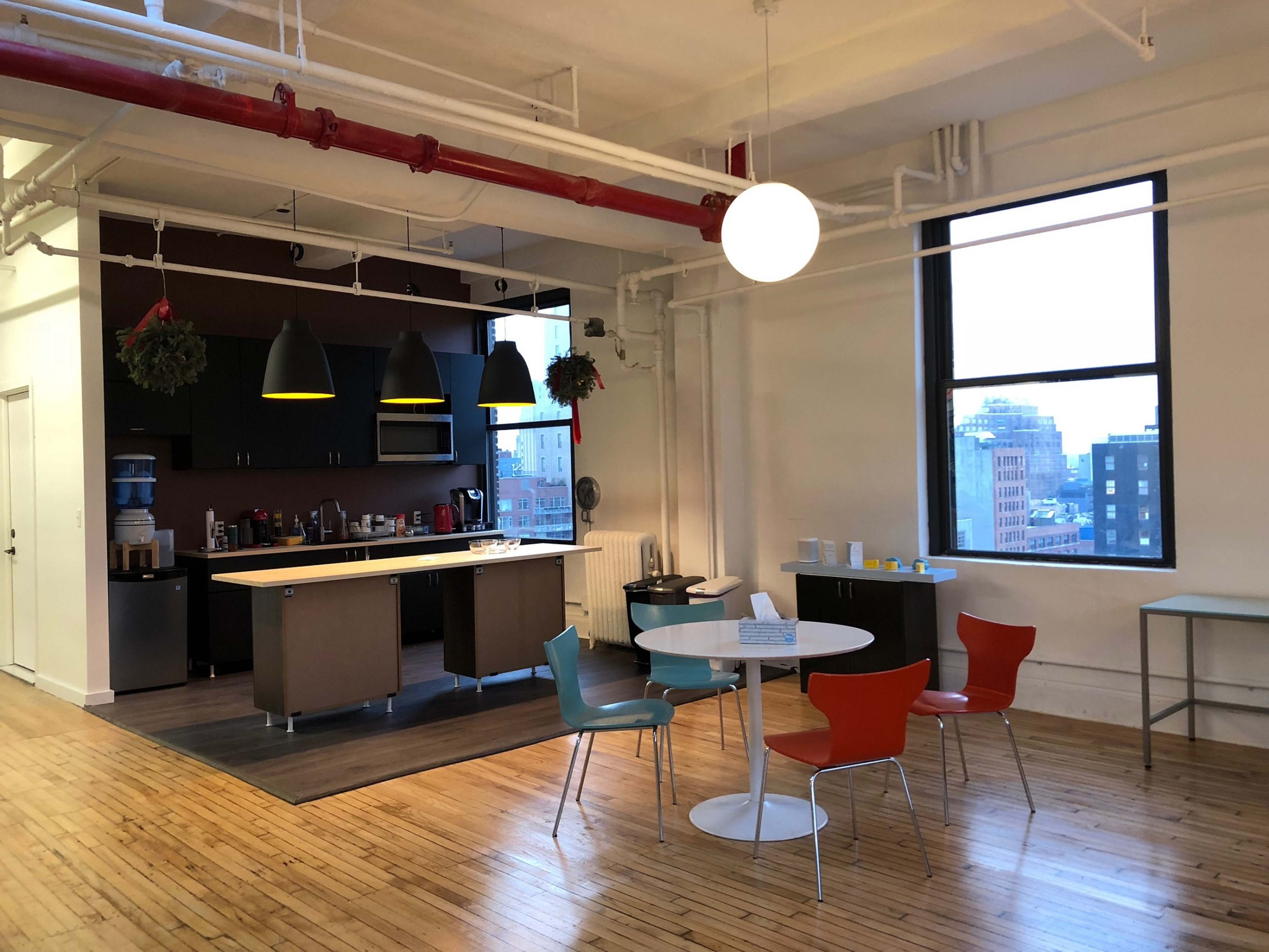 An office break room with a kitchen area, a round table with colorful chairs, and a large window overlooking a cityscape.