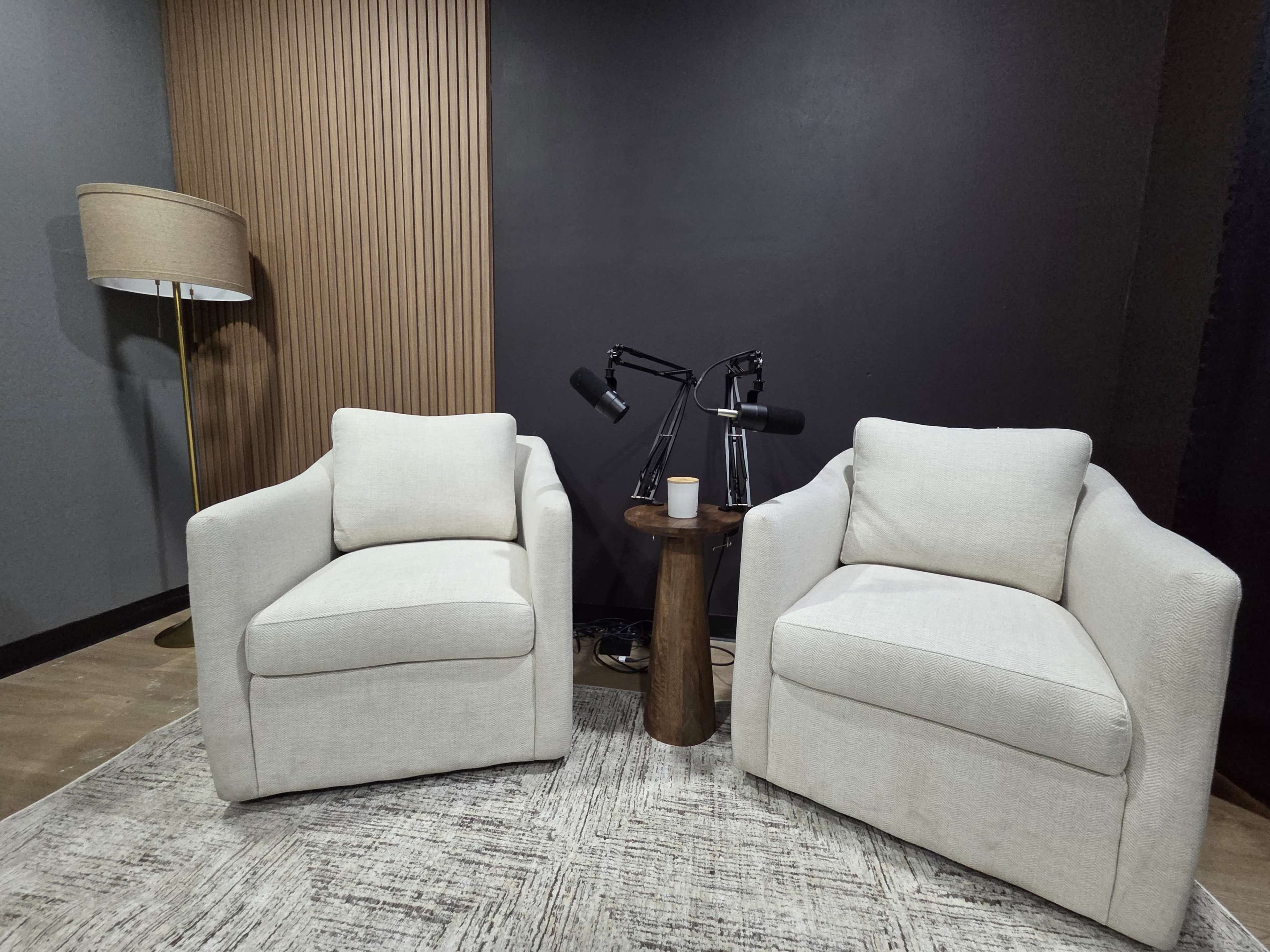 The image shows two white armchairs positioned next to a small wooden table, with a lamp in the background and a dark wall behind.
