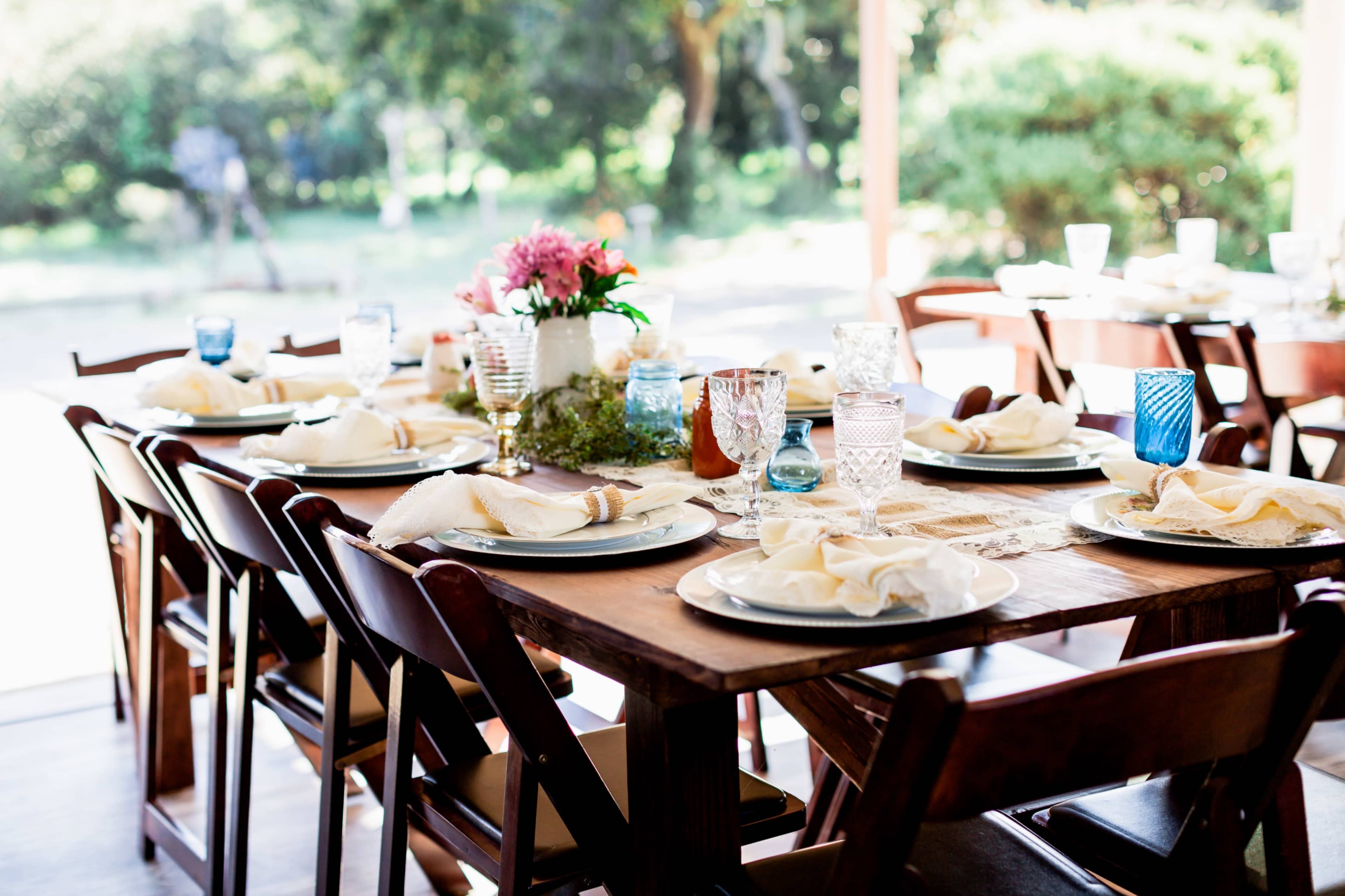 A long wooden dining table is set with plates, napkins, and decorative glassware, surrounded by chairs, in a bright outdoor setting.