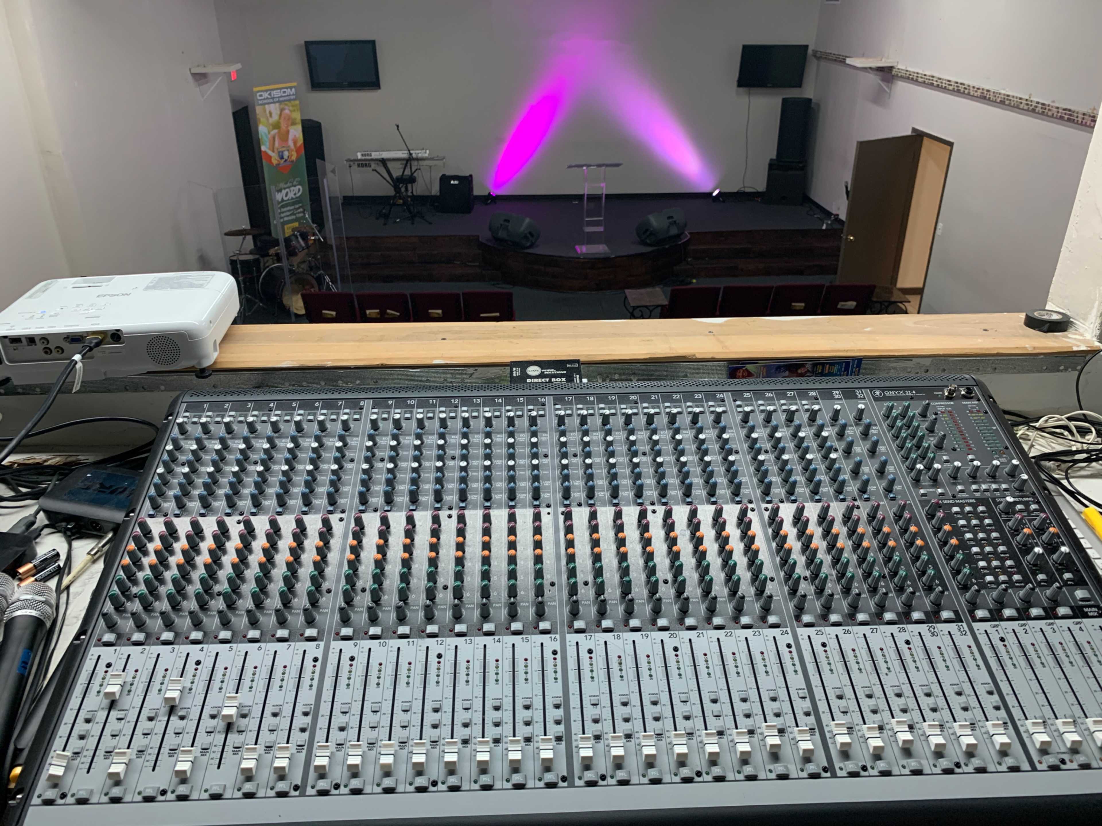 A sound mixing console in the foreground with a stage set up in the background featuring purple lighting and musical instruments.