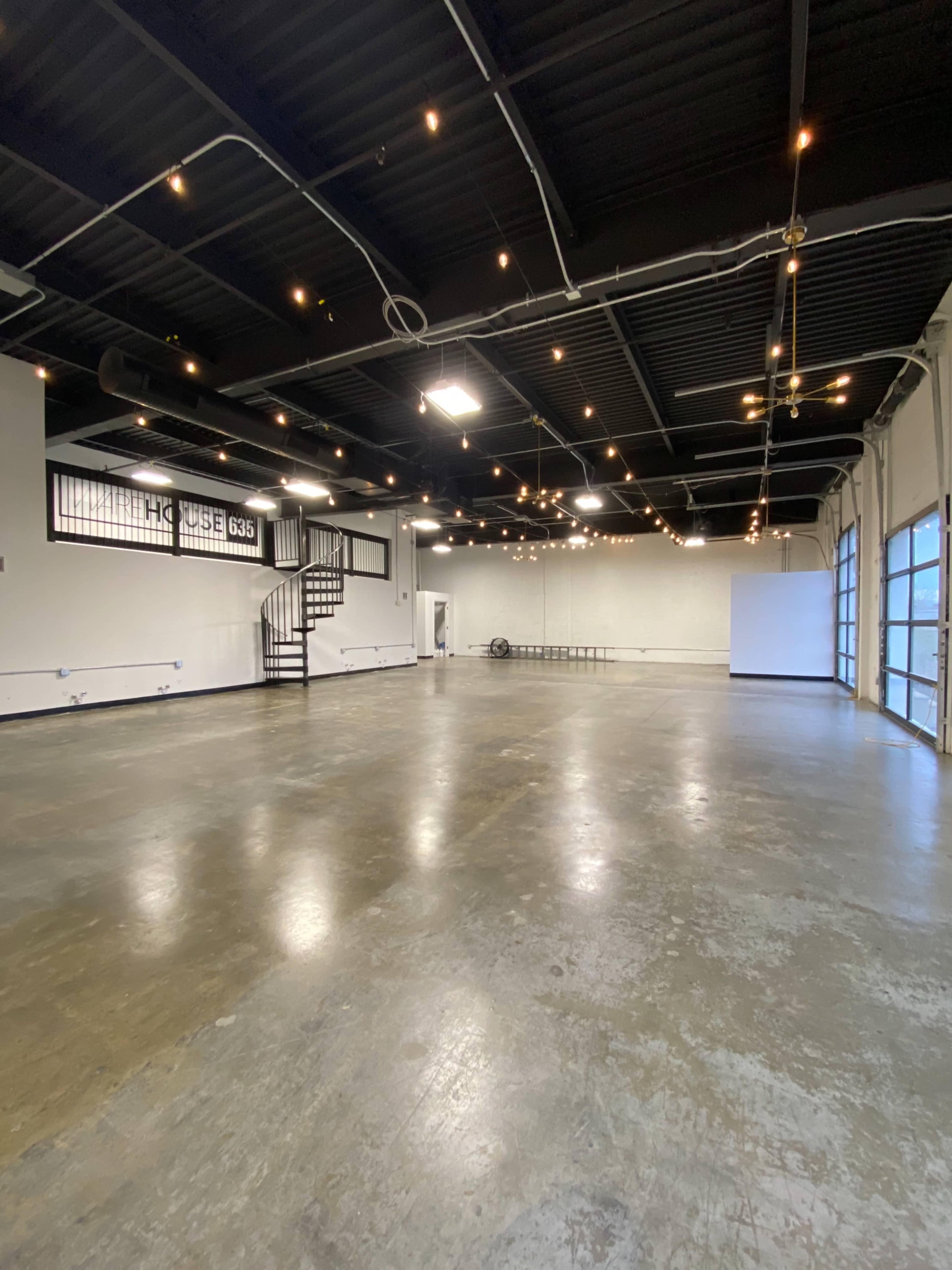 An empty industrial space with concrete flooring, exposed ceiling beams, and string lights overhead.