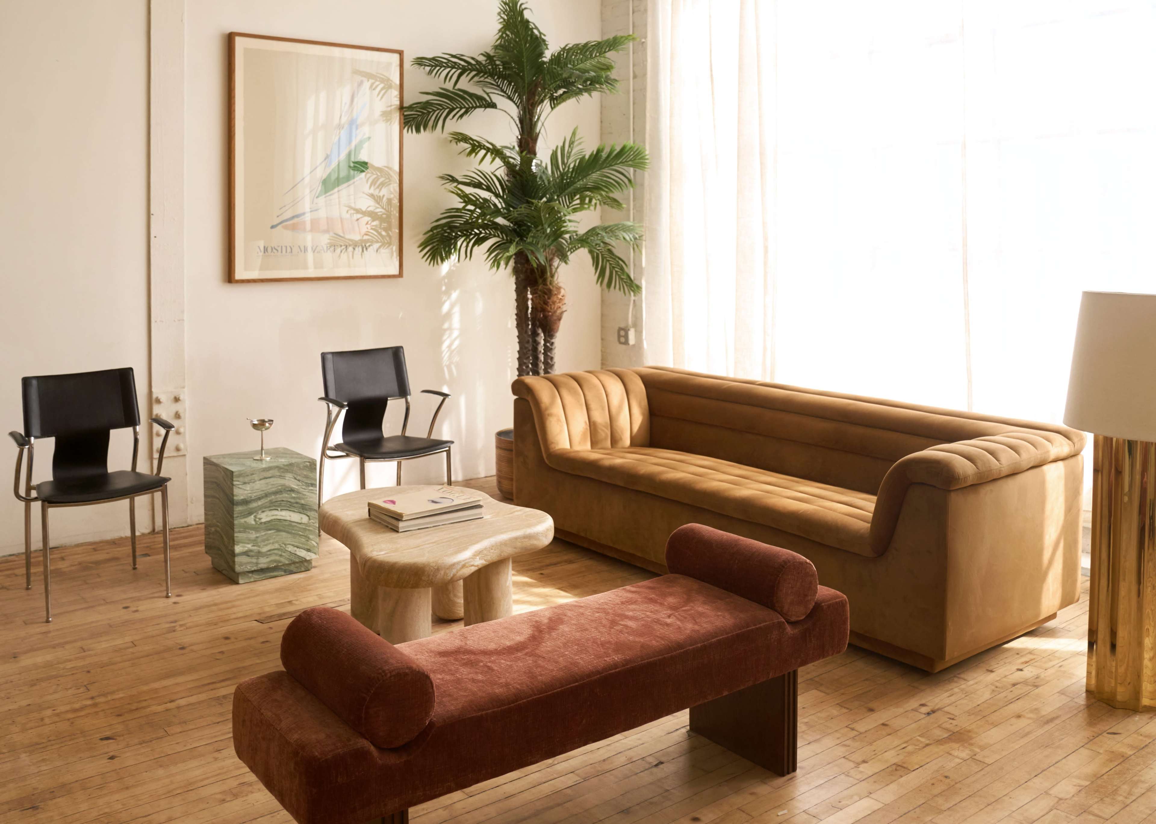 The image features a stylish interior space with a brown sofa, a wooden coffee table, two black chairs, a green marble side table, and a decorative floor lamp, all set against a backdrop of large windows and a potted palm.