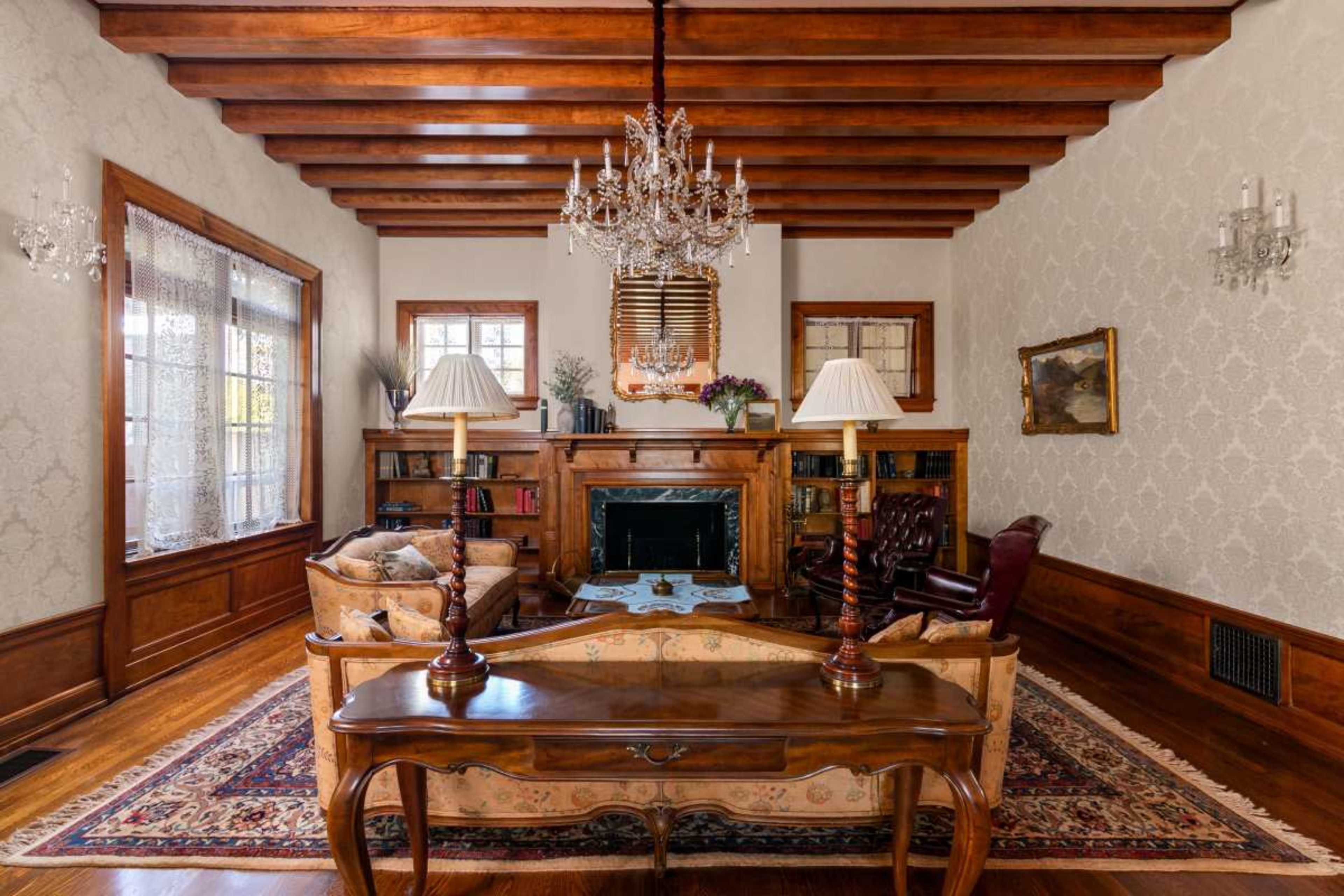 The image shows a classic living room featuring wooden beams on the ceiling, a chandelier, a fireplace, bookshelves, and elegant furniture.