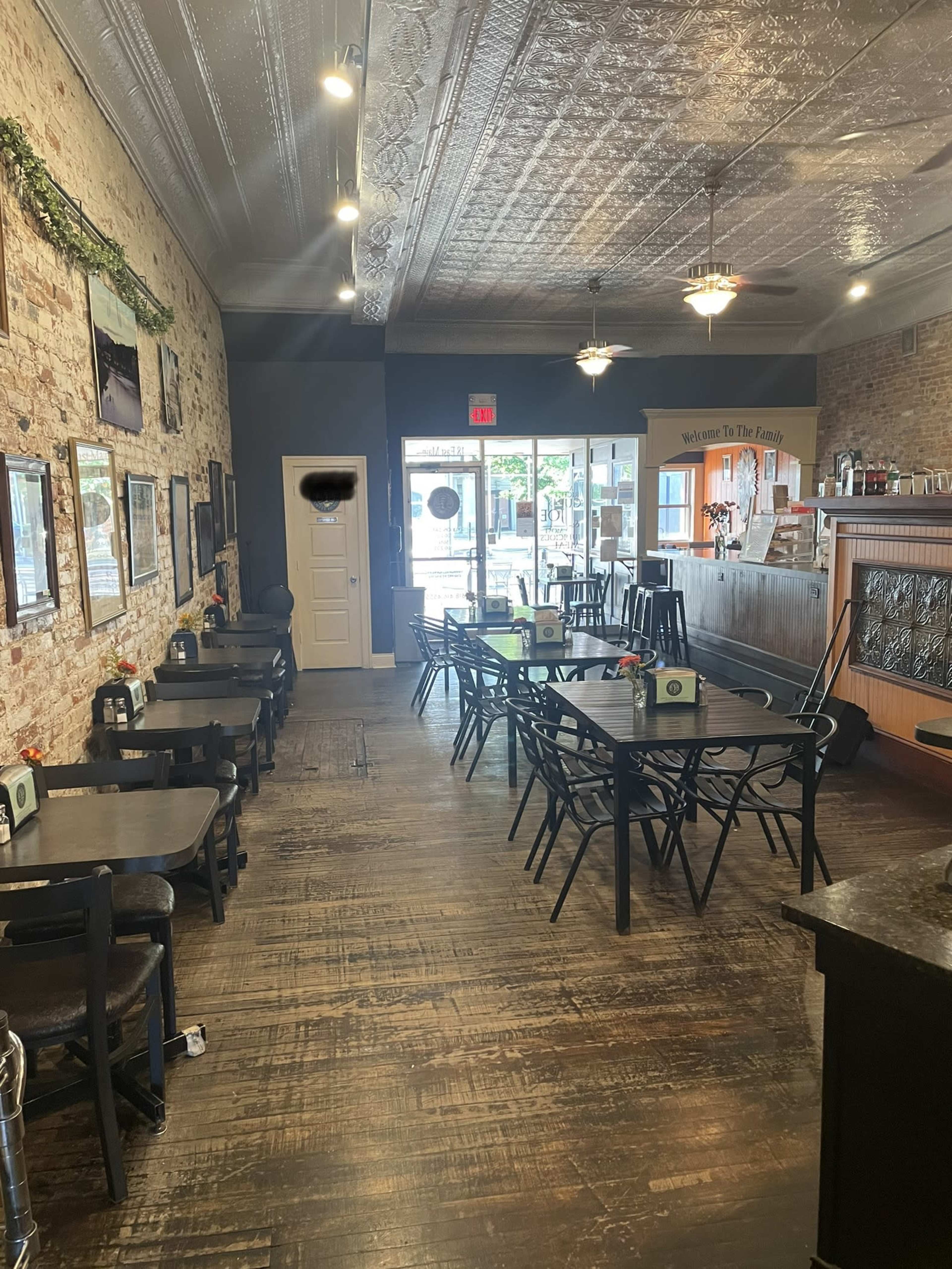 A cozy café interior with wooden flooring, tables and chairs arranged for dining, and exposed brick walls adorned with framed photographs.
