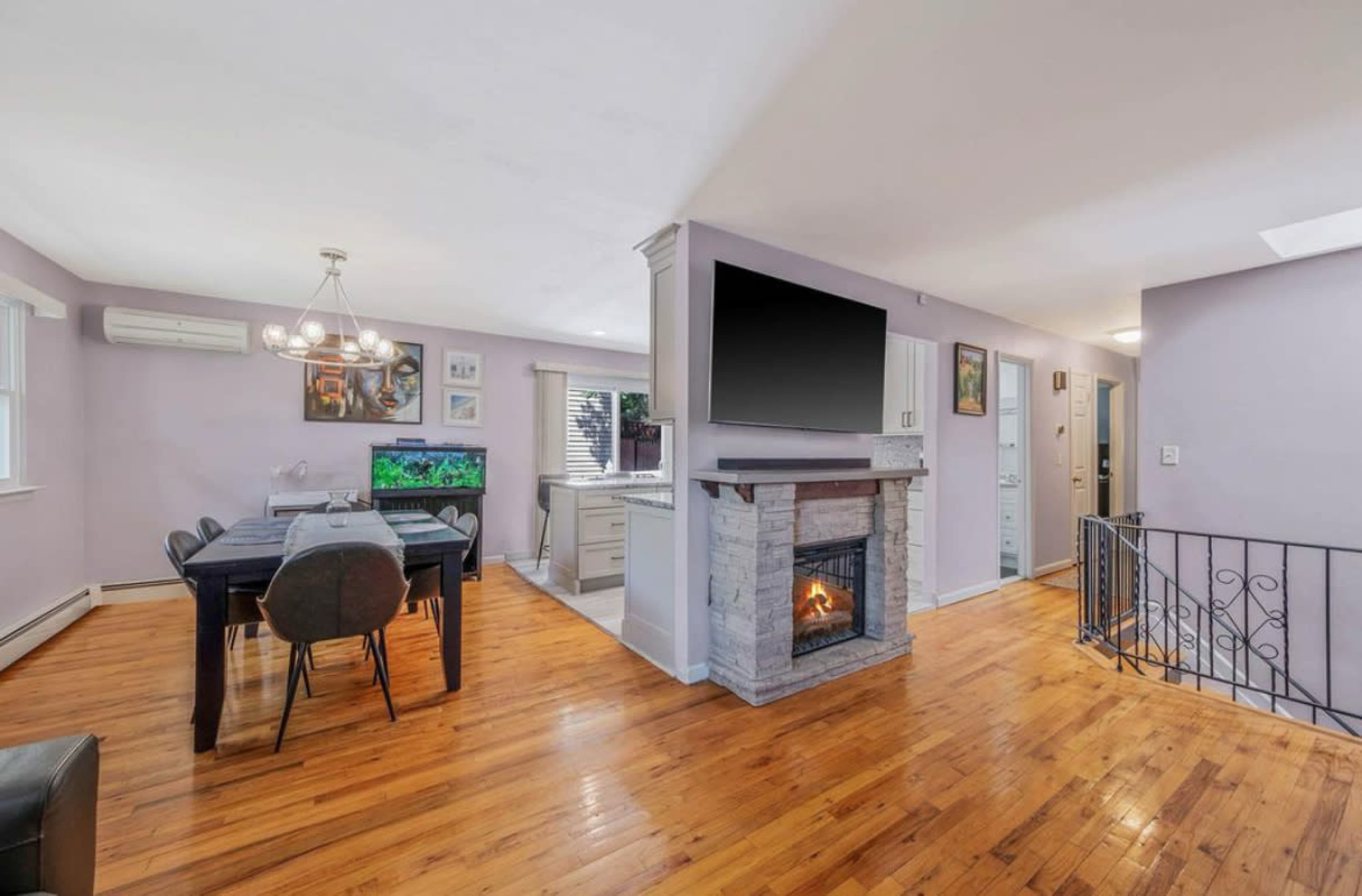 The image shows a living area with a dining table, an electric fireplace, and a wall-mounted television, all featuring light-colored walls and hardwood floors.