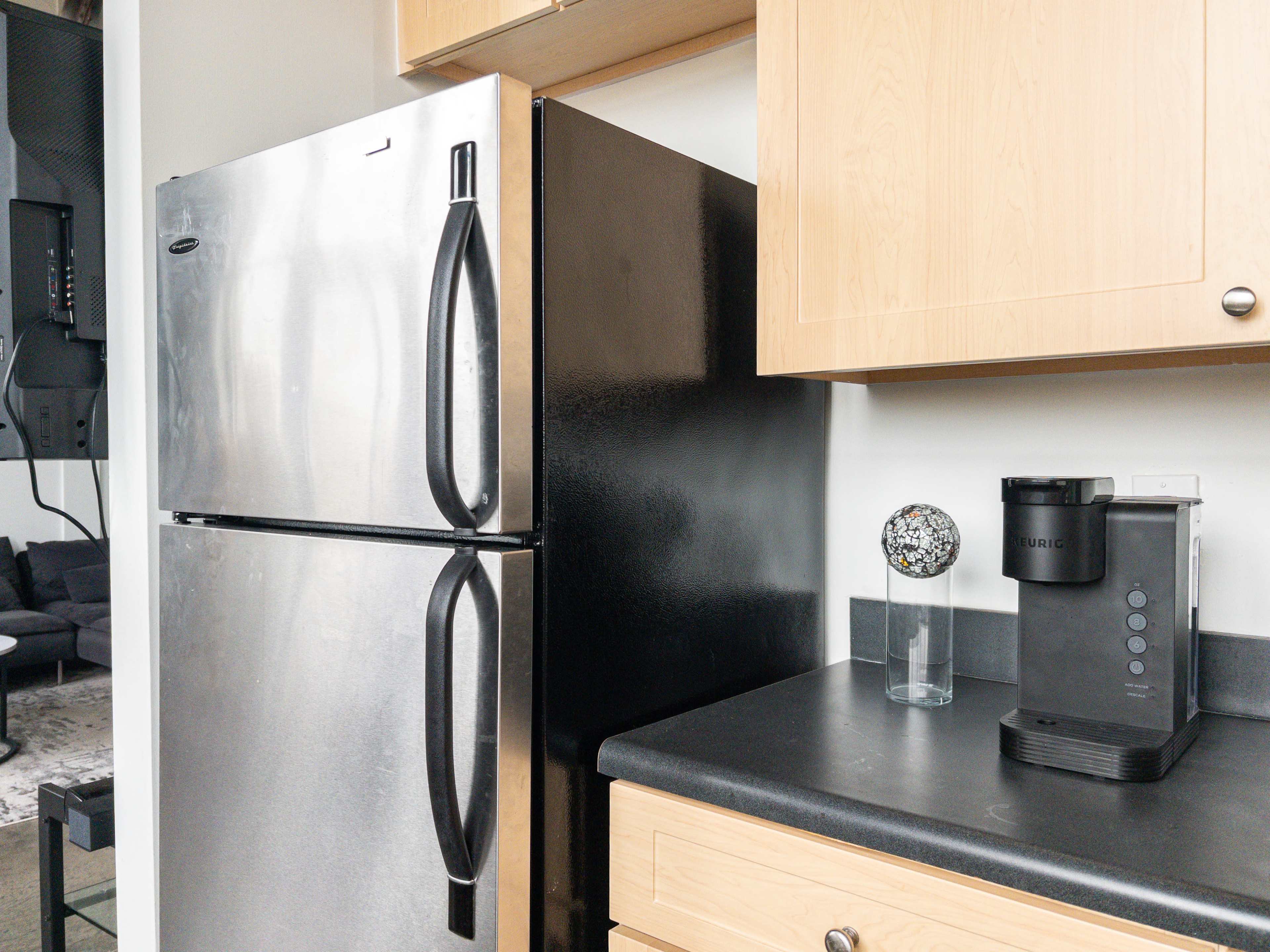 The image shows a stainless steel refrigerator next to a black countertop with a coffee maker and a glass on it.