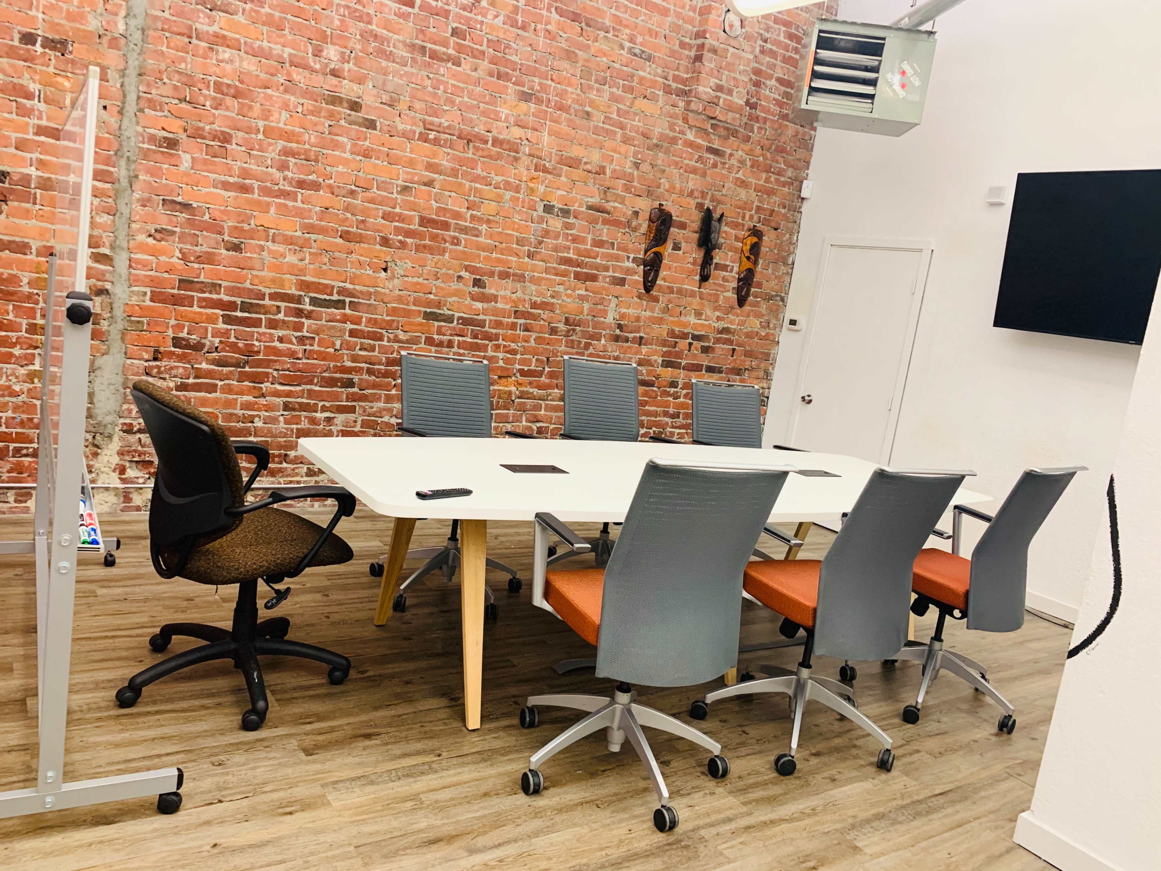 A modern conference room with a long white table surrounded by six office chairs, a brick wall in the background, and a wall-mounted screen.