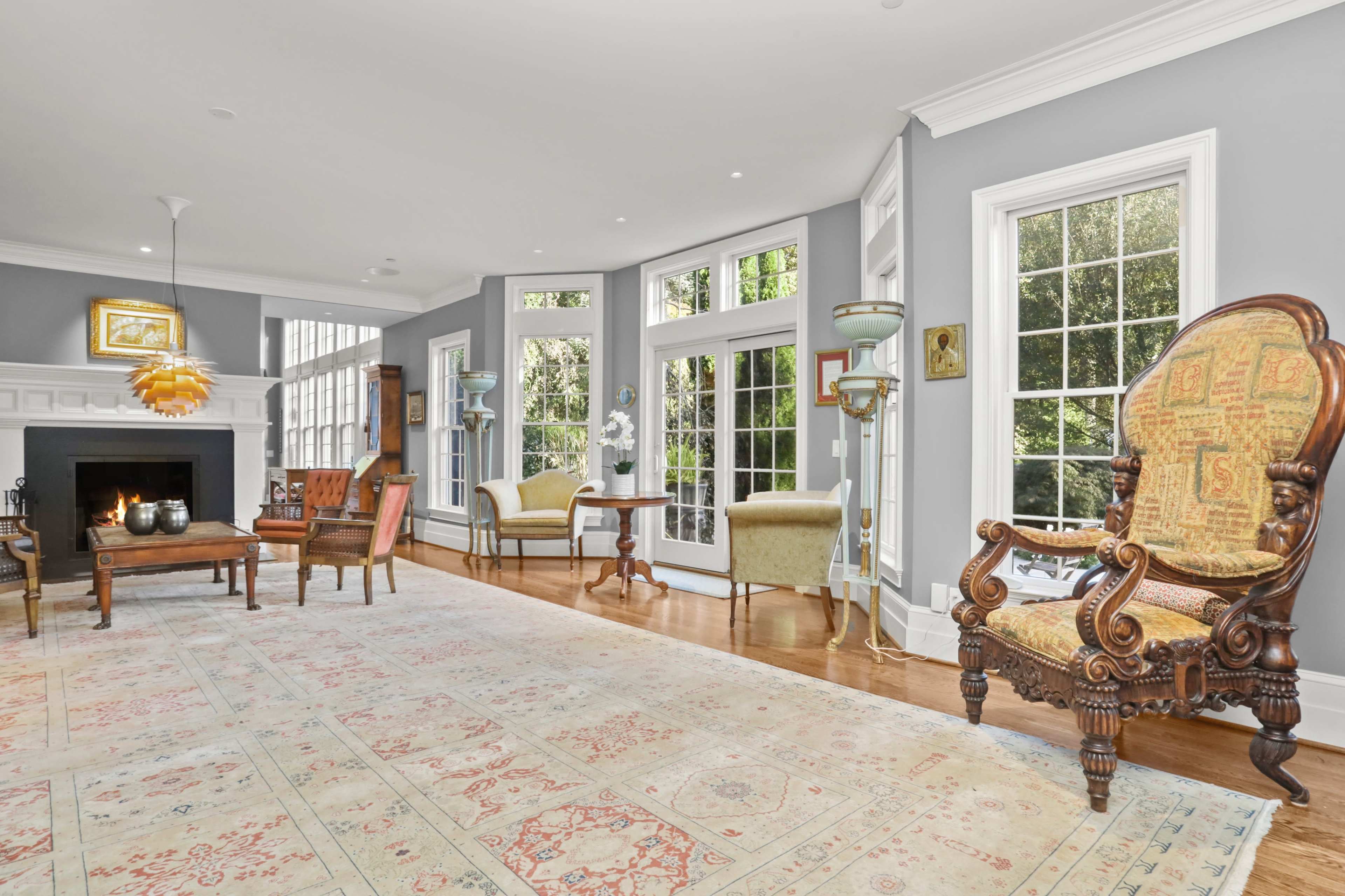 The room features a light-filled living area with large windows, wooden floors, and elegant furniture, including a patterned armchair and a fireplace.