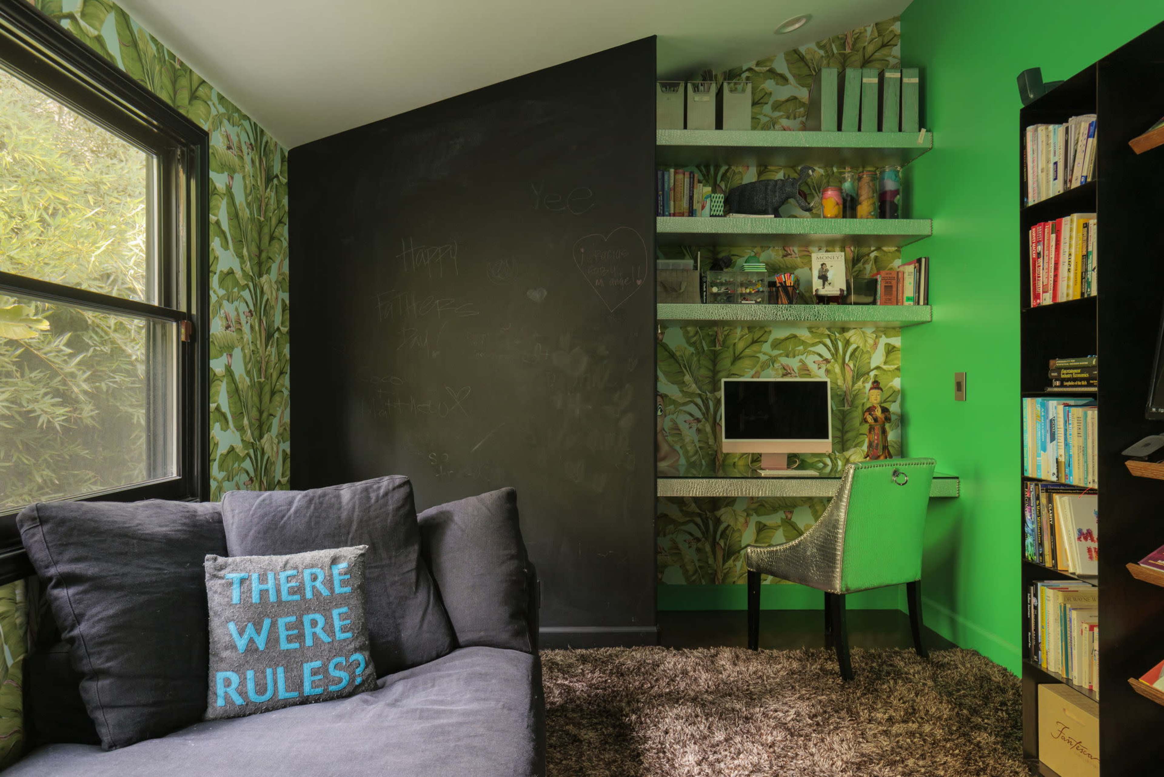 The image shows a small office space with green walls, a black chalkboard, a gray couch, an ergonomic chair, and a desk with an Apple computer.