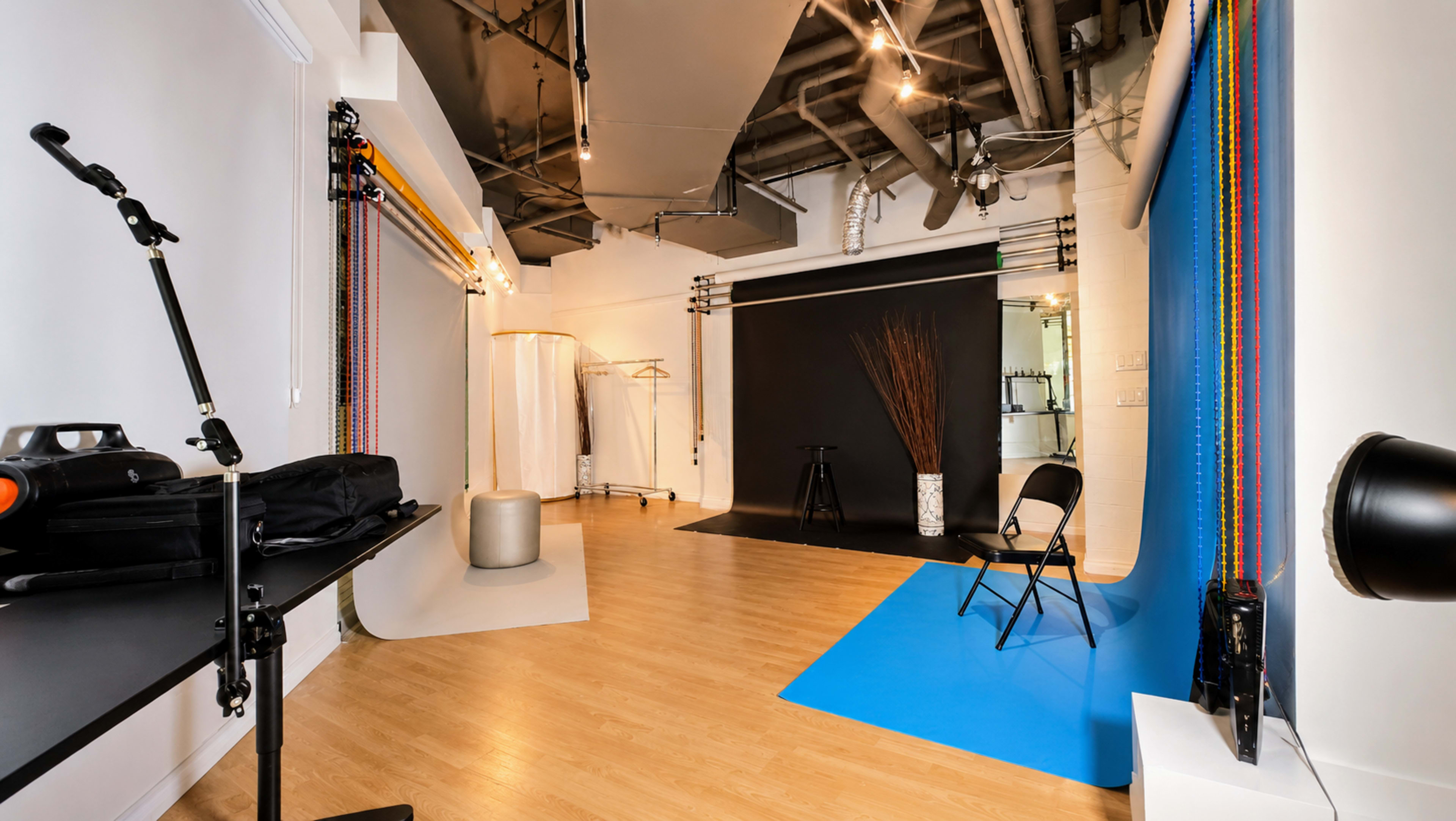 The image shows a photography studio featuring a black backdrop, blue backdrop, a chair, a clothing rack, and various lighting equipment in a well-lit space with wooden flooring.