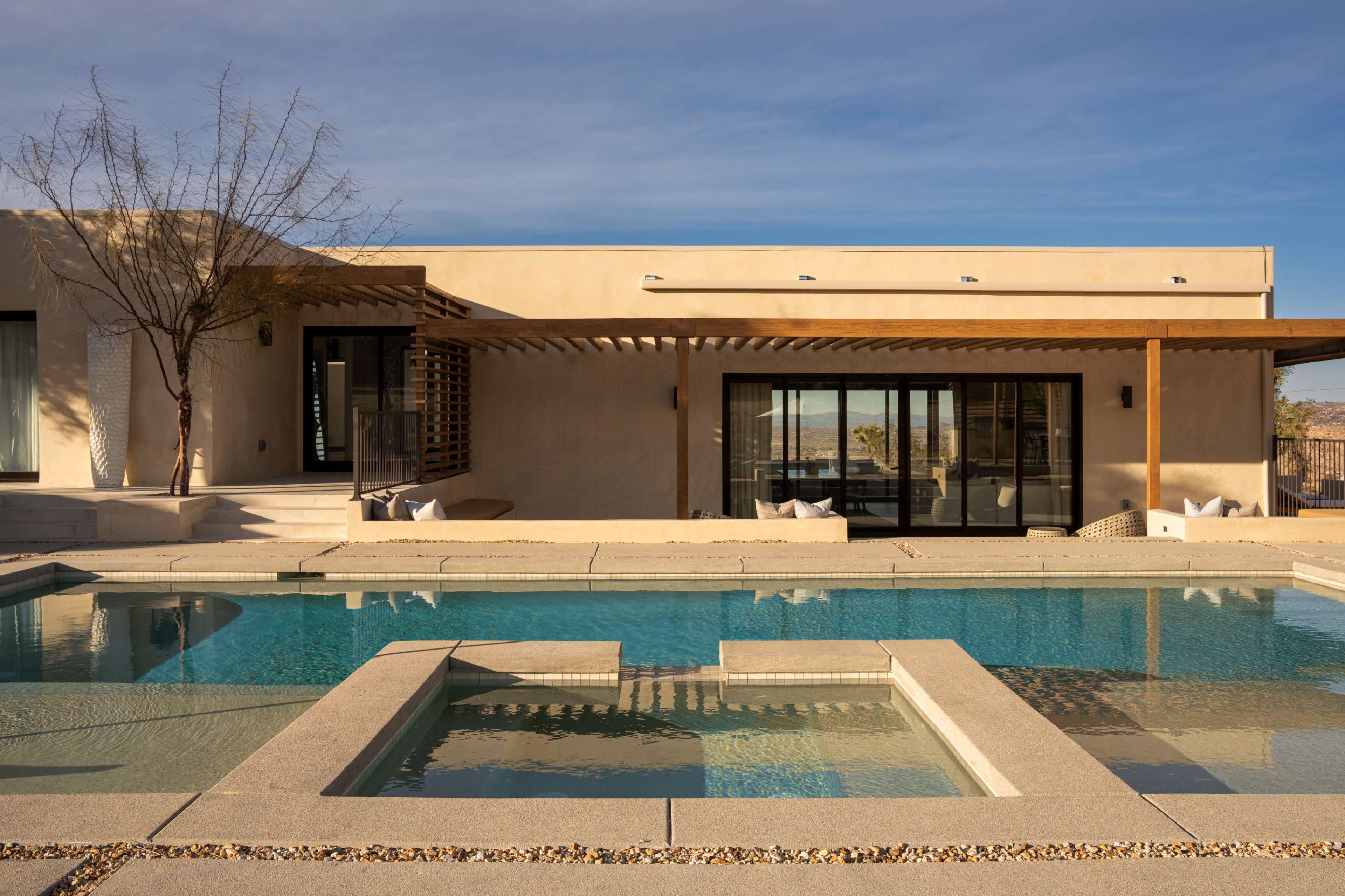 A modern desert house features a clear blue pool surrounded by a stone patio and minimal landscaping.