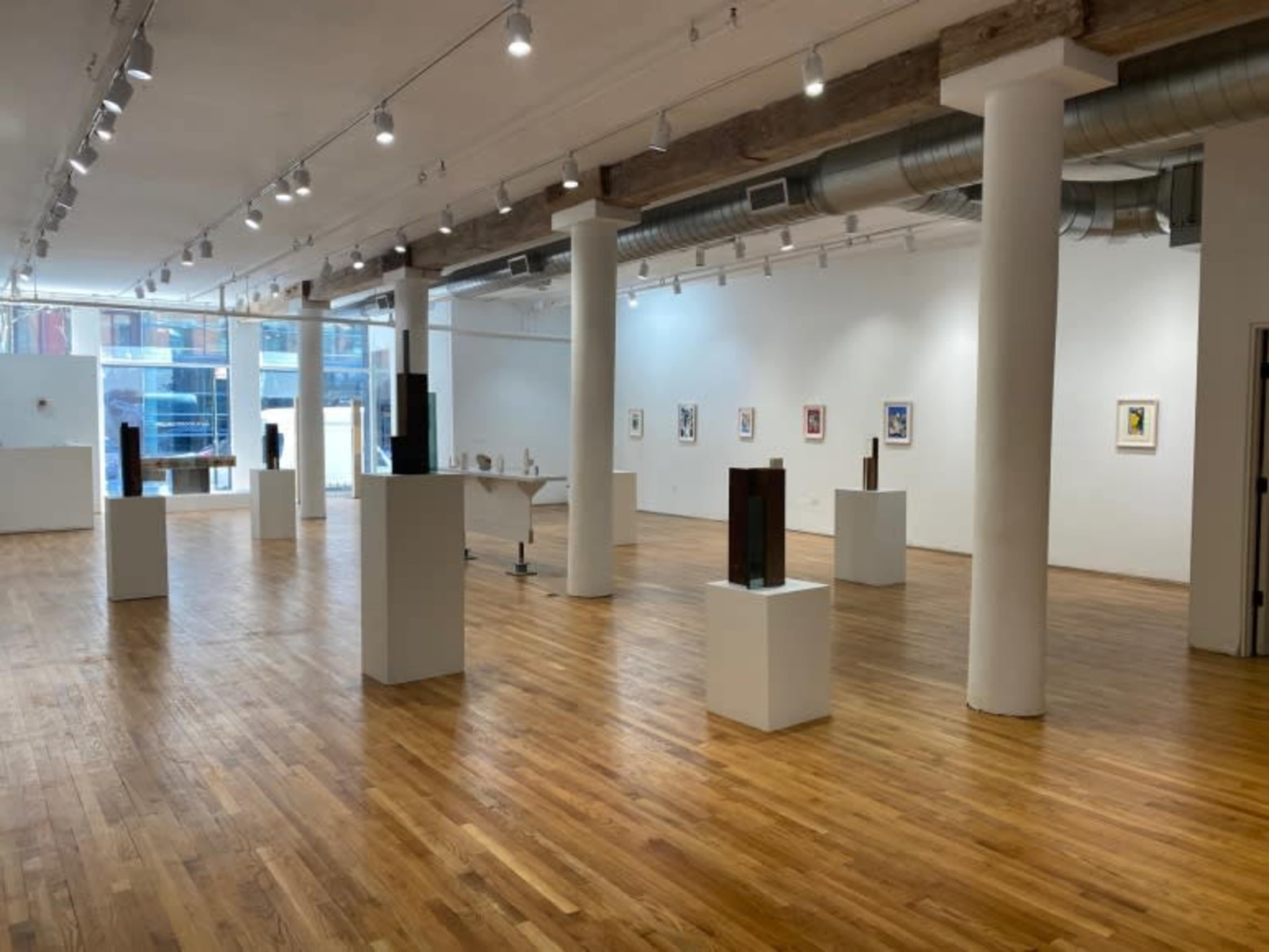 An art gallery with wooden flooring, several sculptures on white pedestals, and framed artworks hanging on the walls.