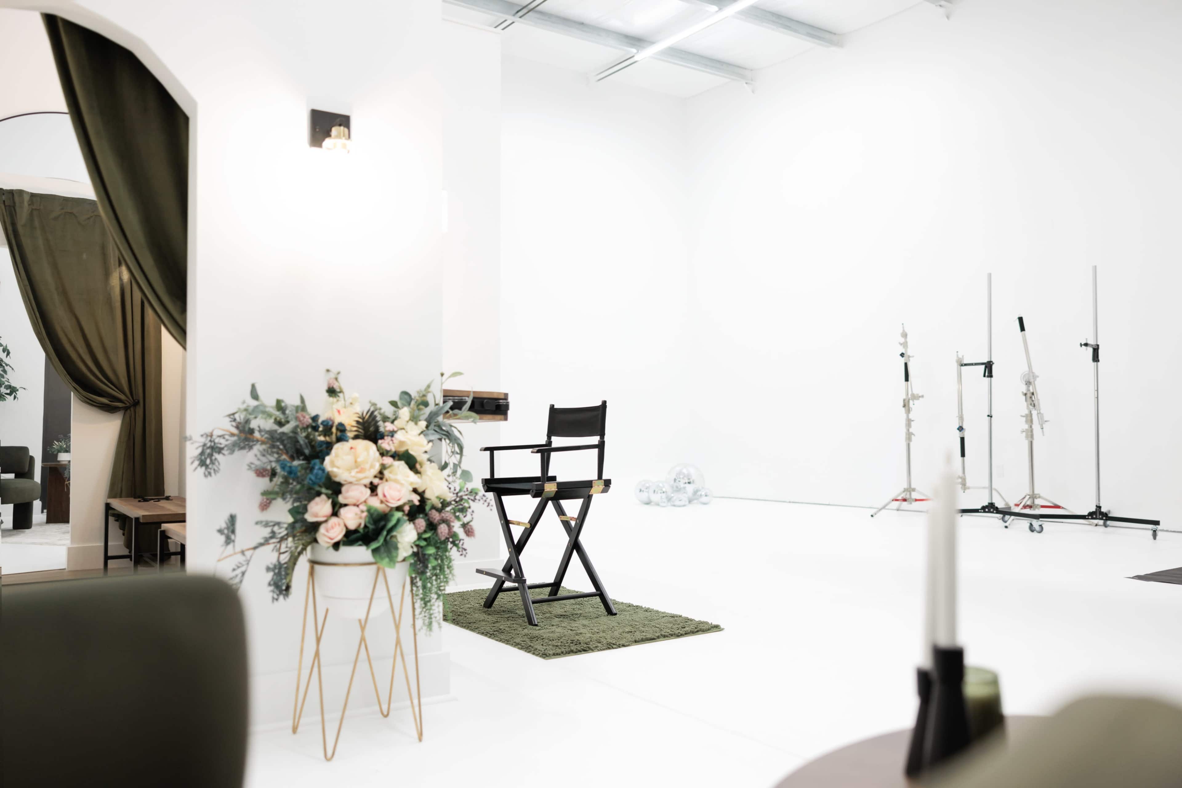 The image shows a bright, white studio space featuring a director's chair on a green mat, a stylish floral arrangement in a planter, and lighting equipment in the background.