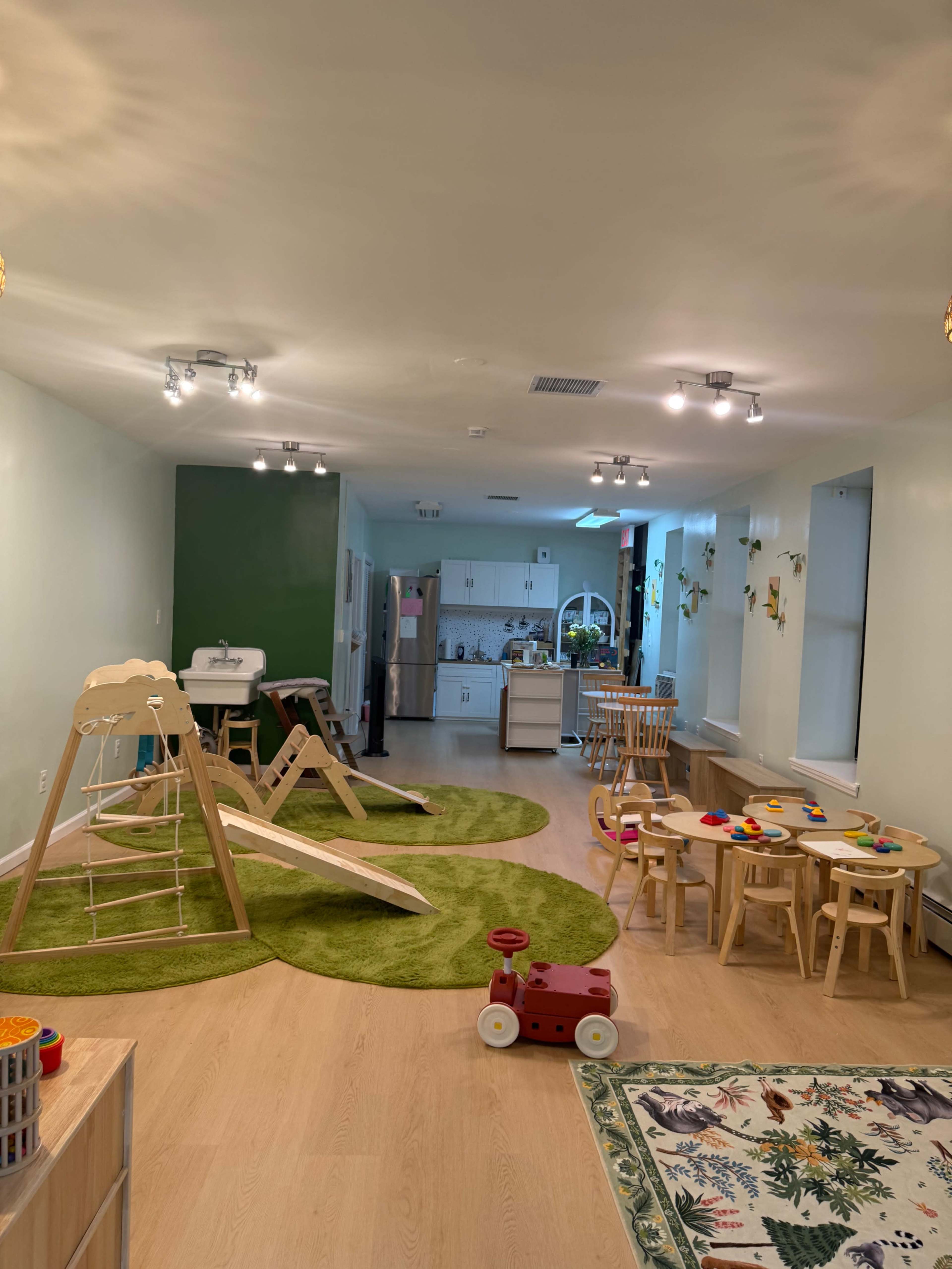The image shows a brightly lit playroom featuring a green carpet, wooden play equipment, a kitchen area in the background, and a variety of toys scattered across a circular table.