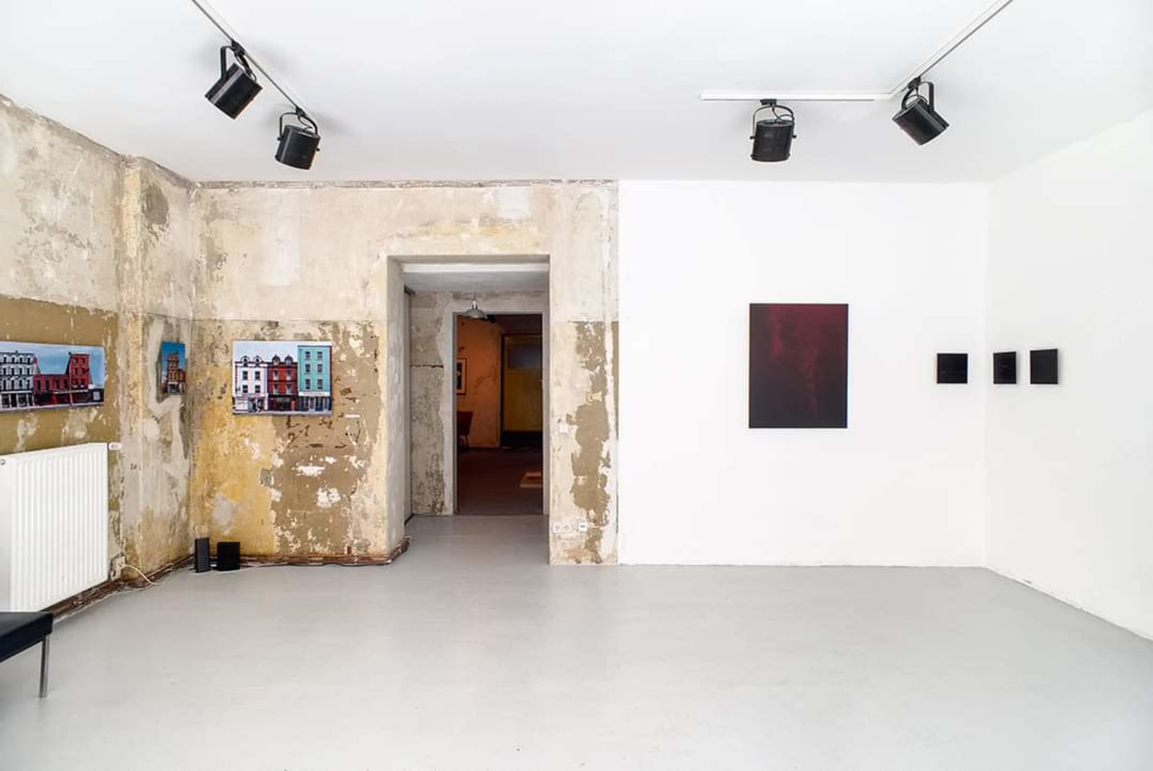 The image shows an art gallery with peeling plaster walls, exhibiting various artworks, including framed pieces on the left and a dark canvas on the right.