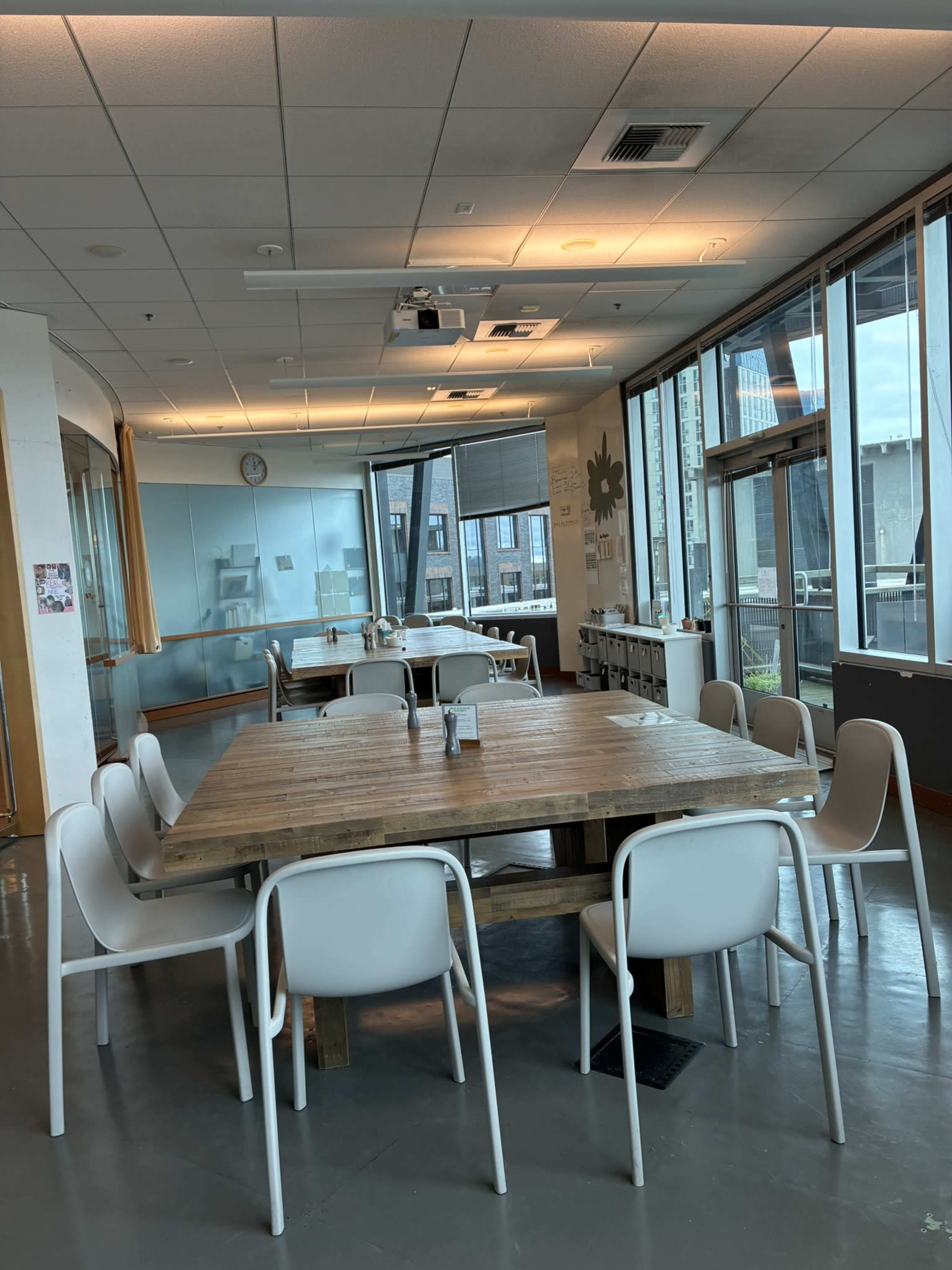 A large wooden table surrounded by white chairs is set in a well-lit room with large windows and a clock on the wall.