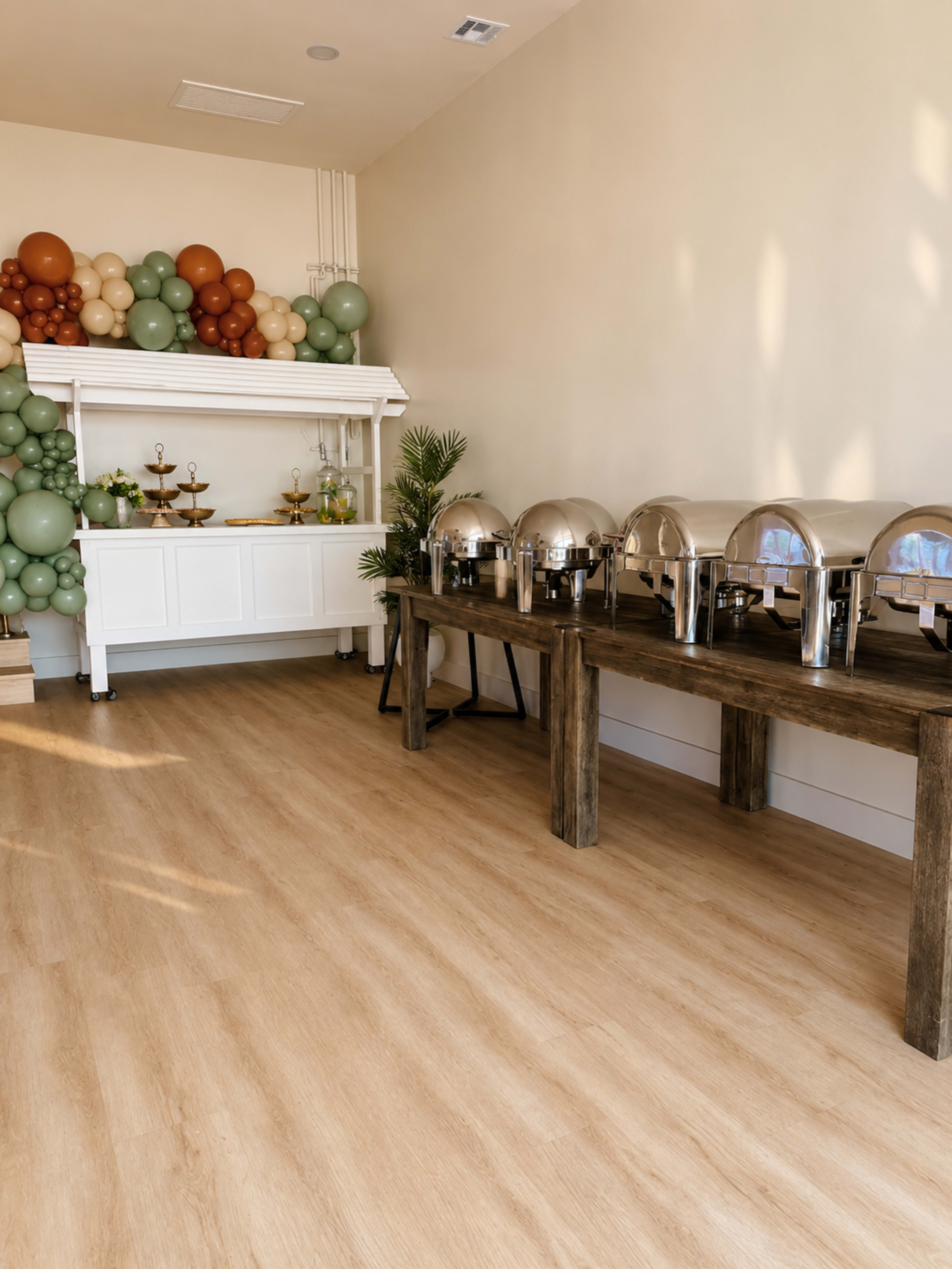 The image shows a banquet setup with several silver chafing dishes on a wooden table, complemented by a backdrop of colorful balloons against a light-colored wall.