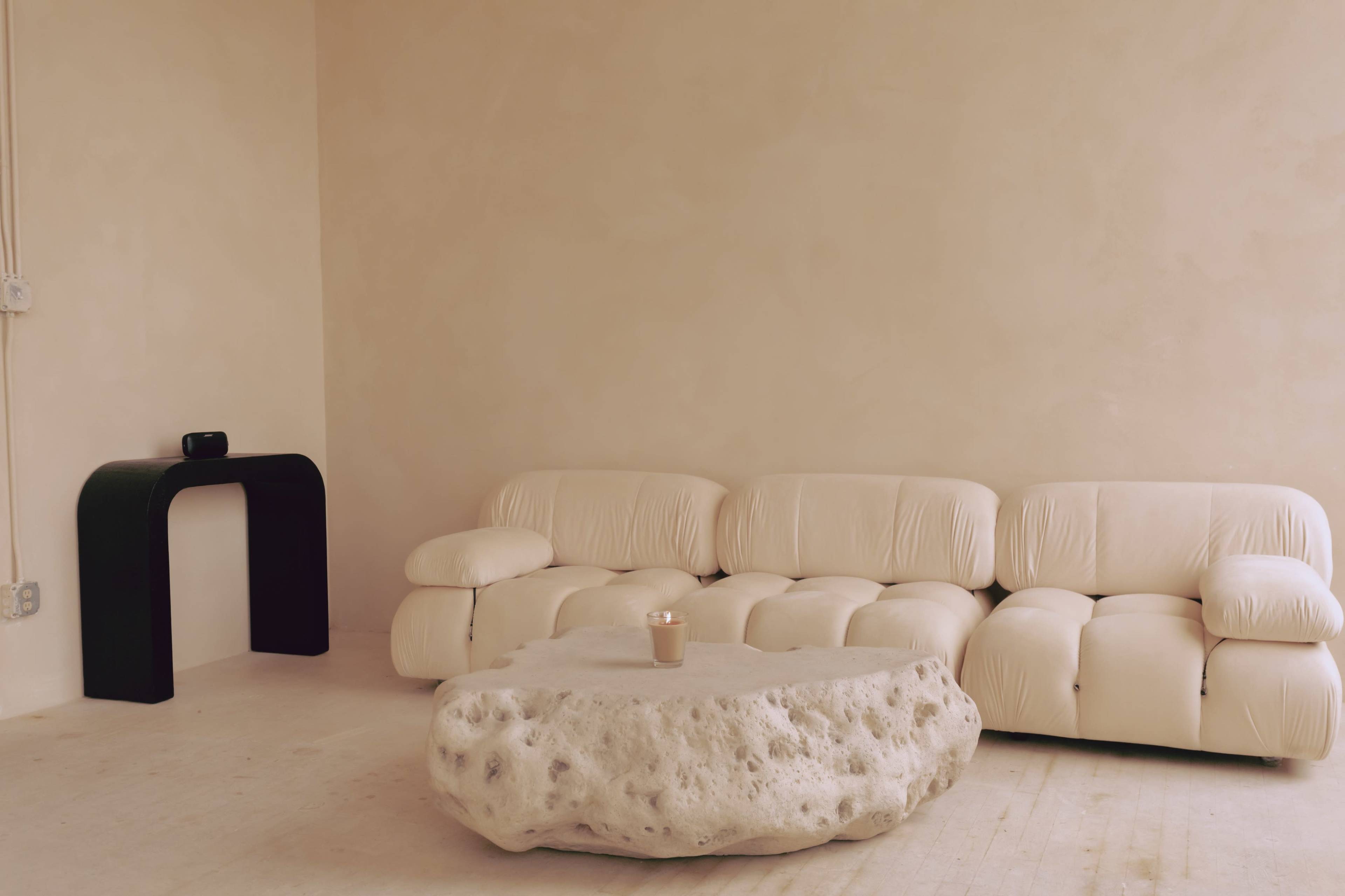 A minimalist living space featuring a beige sectional sofa, a round stone coffee table, and a black archway side table.