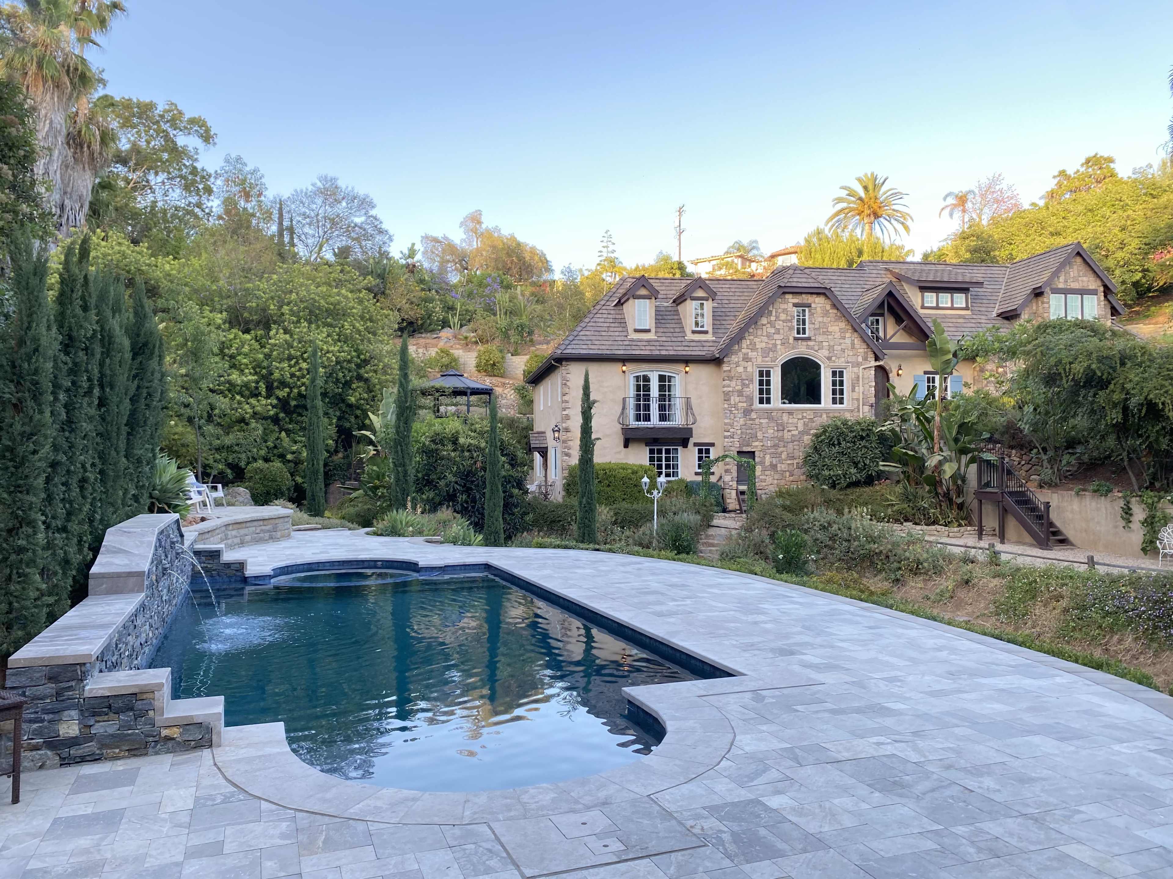A large stone house next to a rectangular swimming pool, surrounded by trees and landscaping.