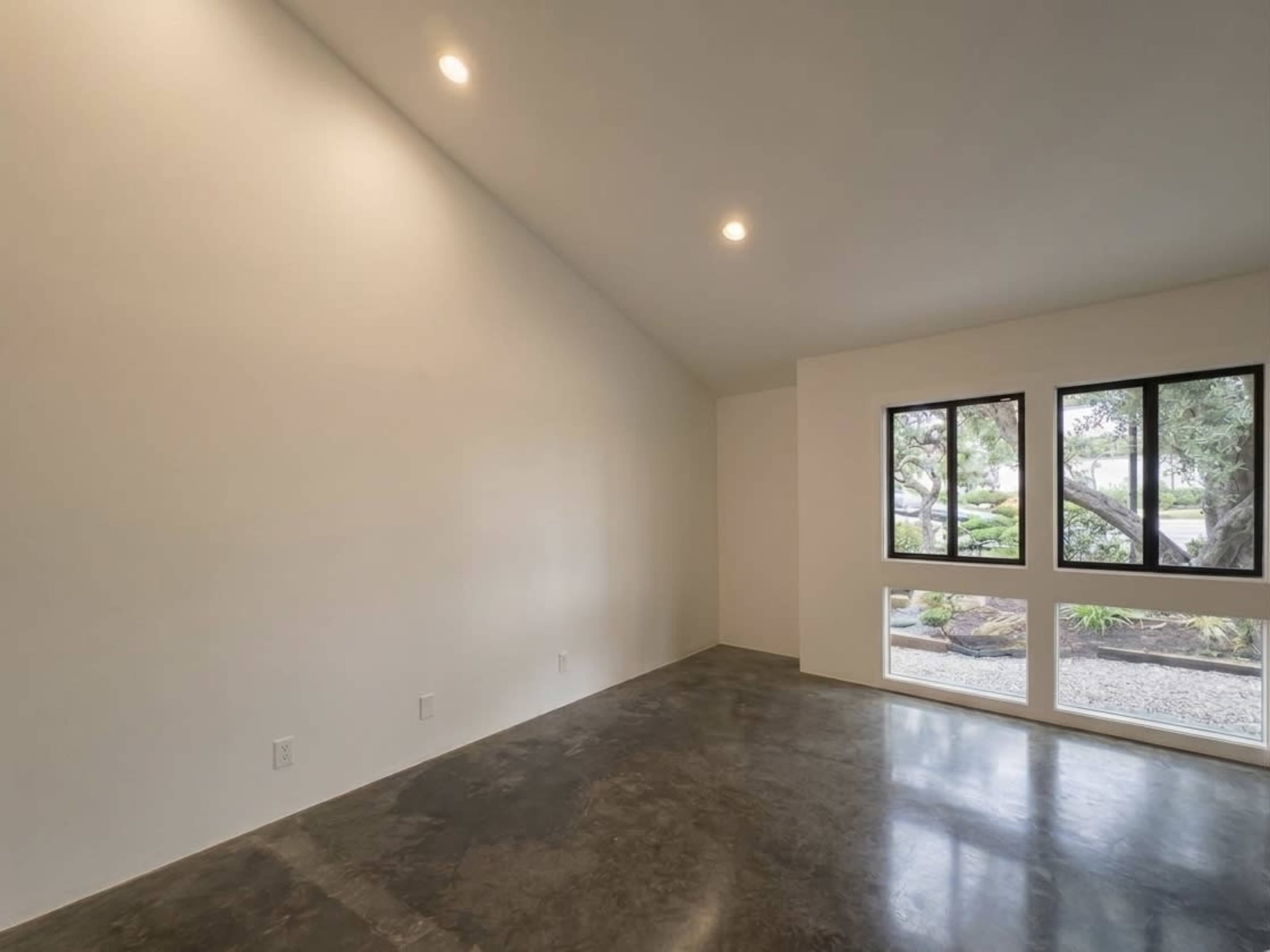 The image shows an empty room with a high ceiling, polished concrete flooring, and two large windows allowing natural light to enter.