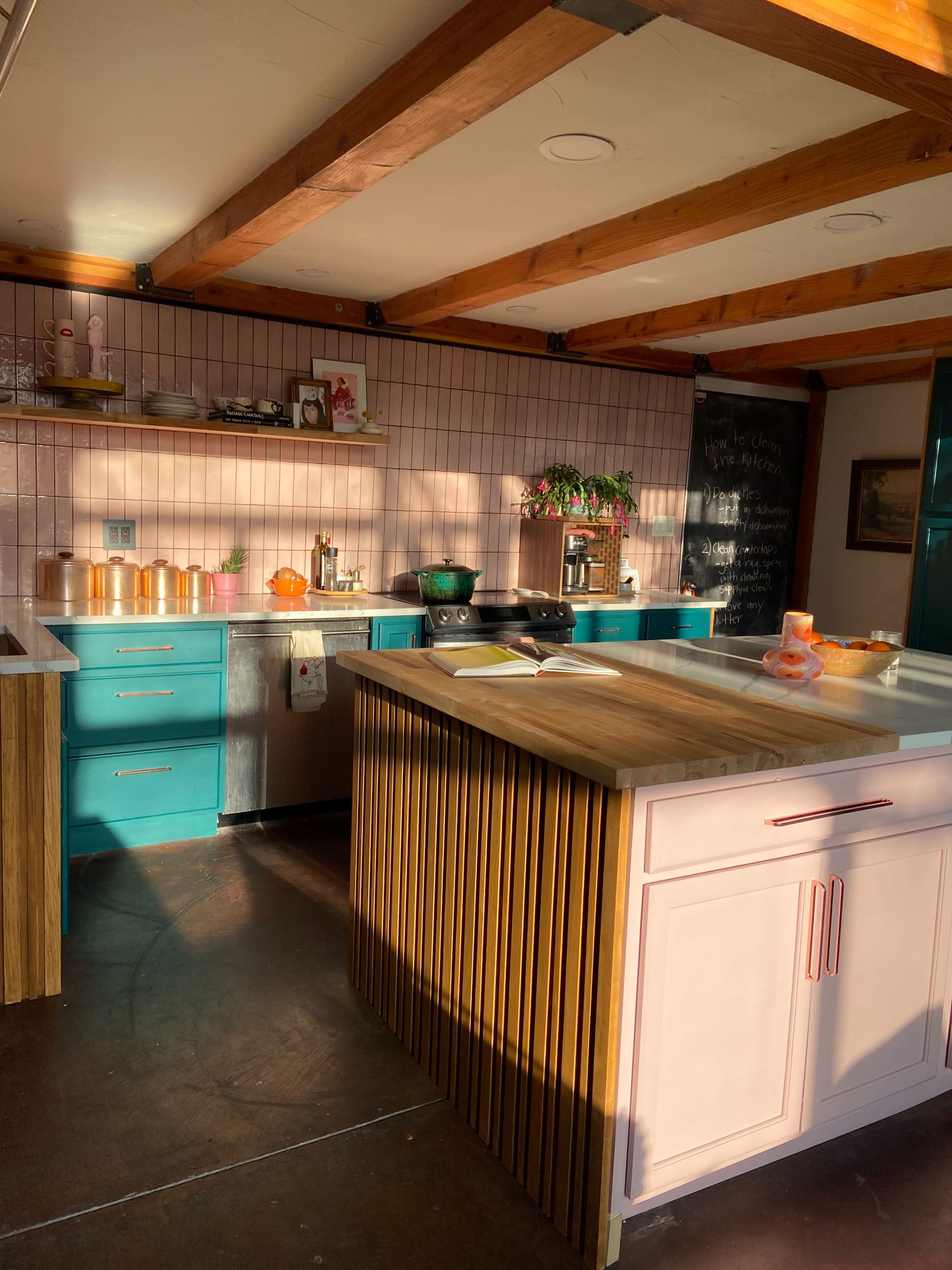 A well-lit kitchen features teal cabinets, a wooden island with vertical slats, and a wall adorned with chalkboard art.