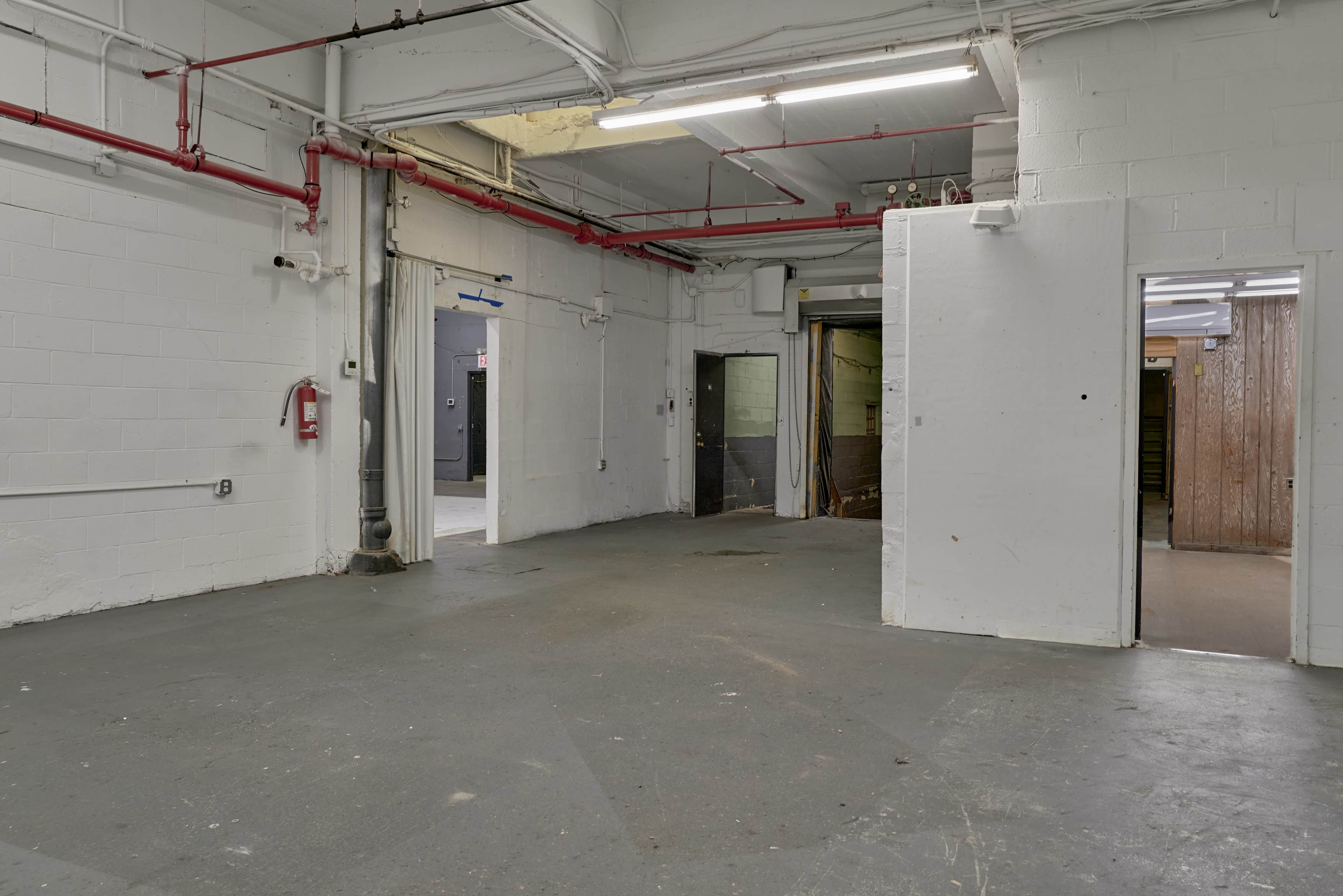 The image shows a large, empty industrial room with concrete floors, white walls, and exposed red piping.