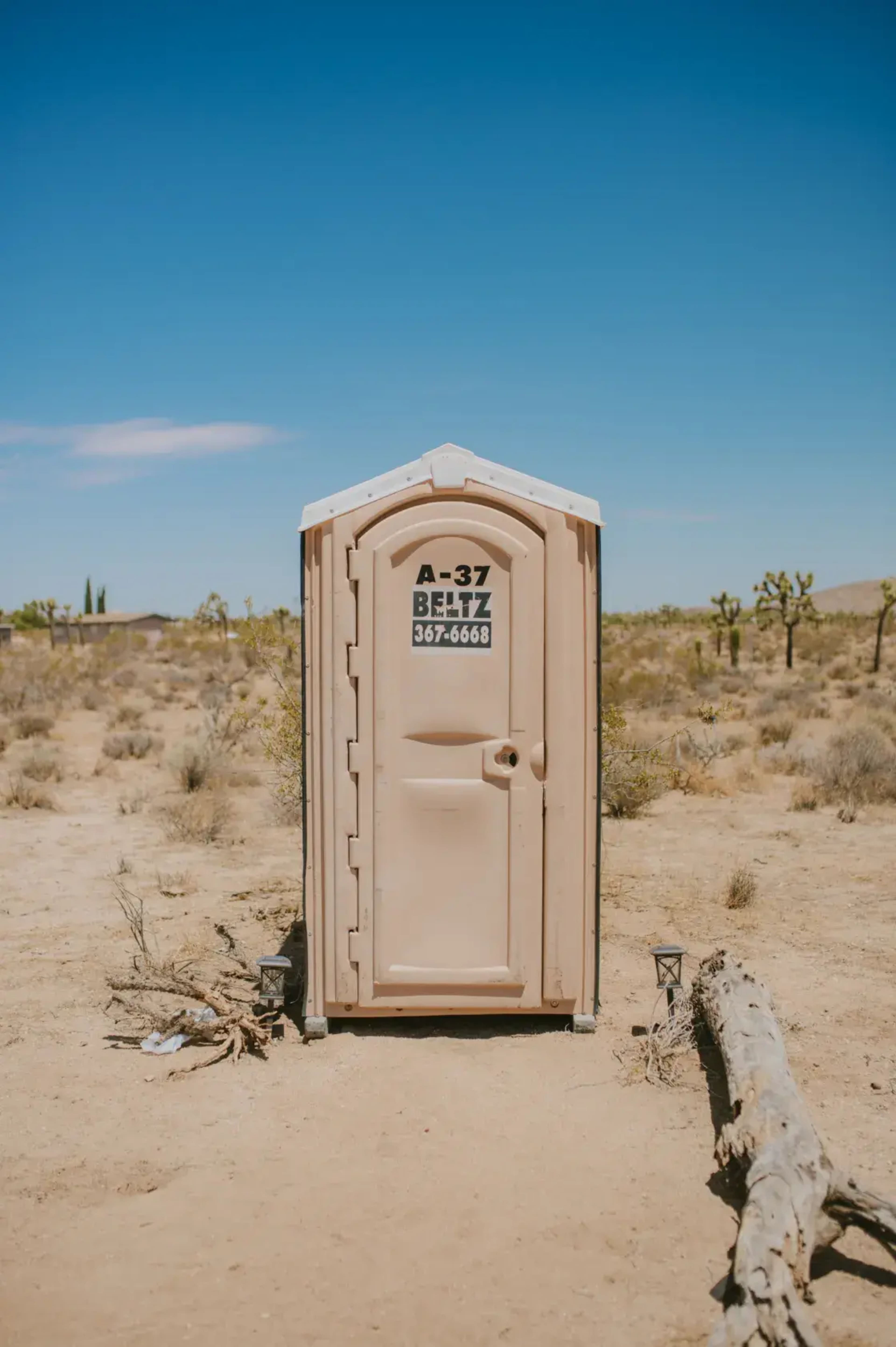A portable restroom is positioned in a desert landscape, surrounded by sparse vegetation and a clear blue sky.
