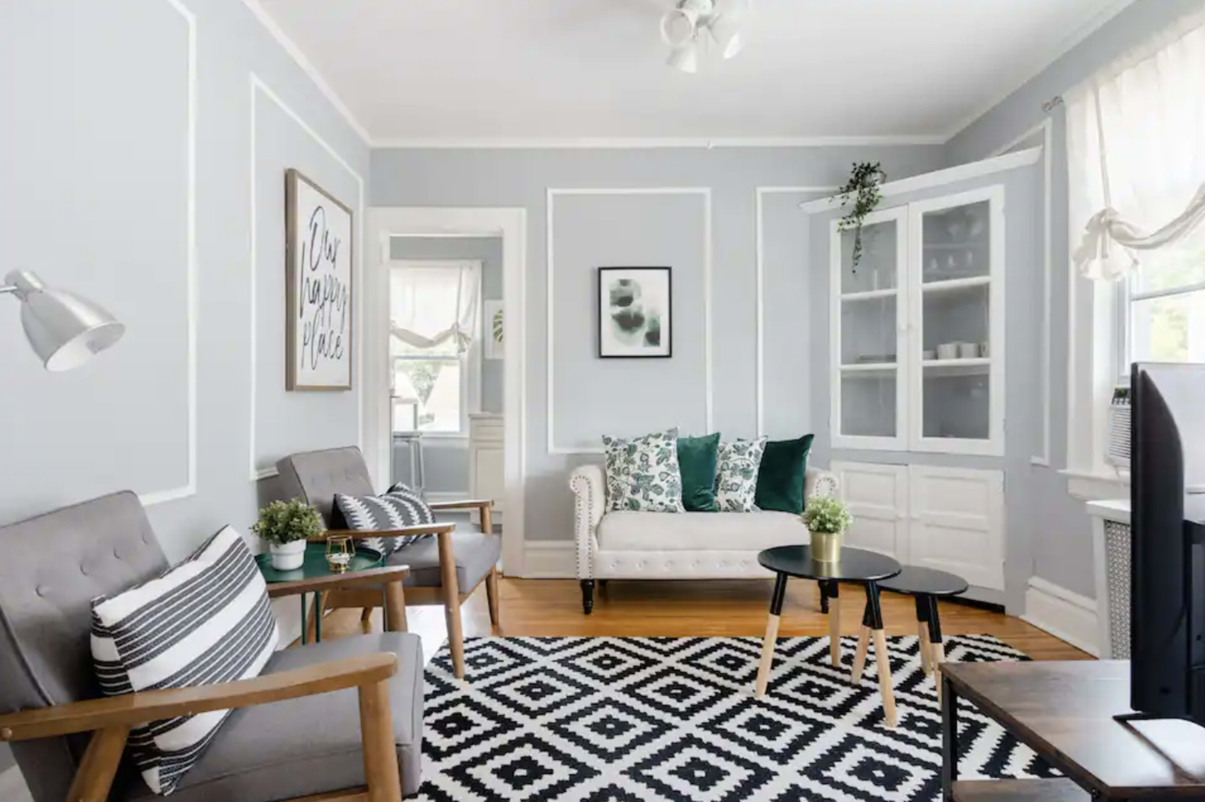 The image shows a living room featuring a light gray color scheme, a striped black and white area rug, a white sofa with green cushions, and two wooden chairs beside a small black coffee table.