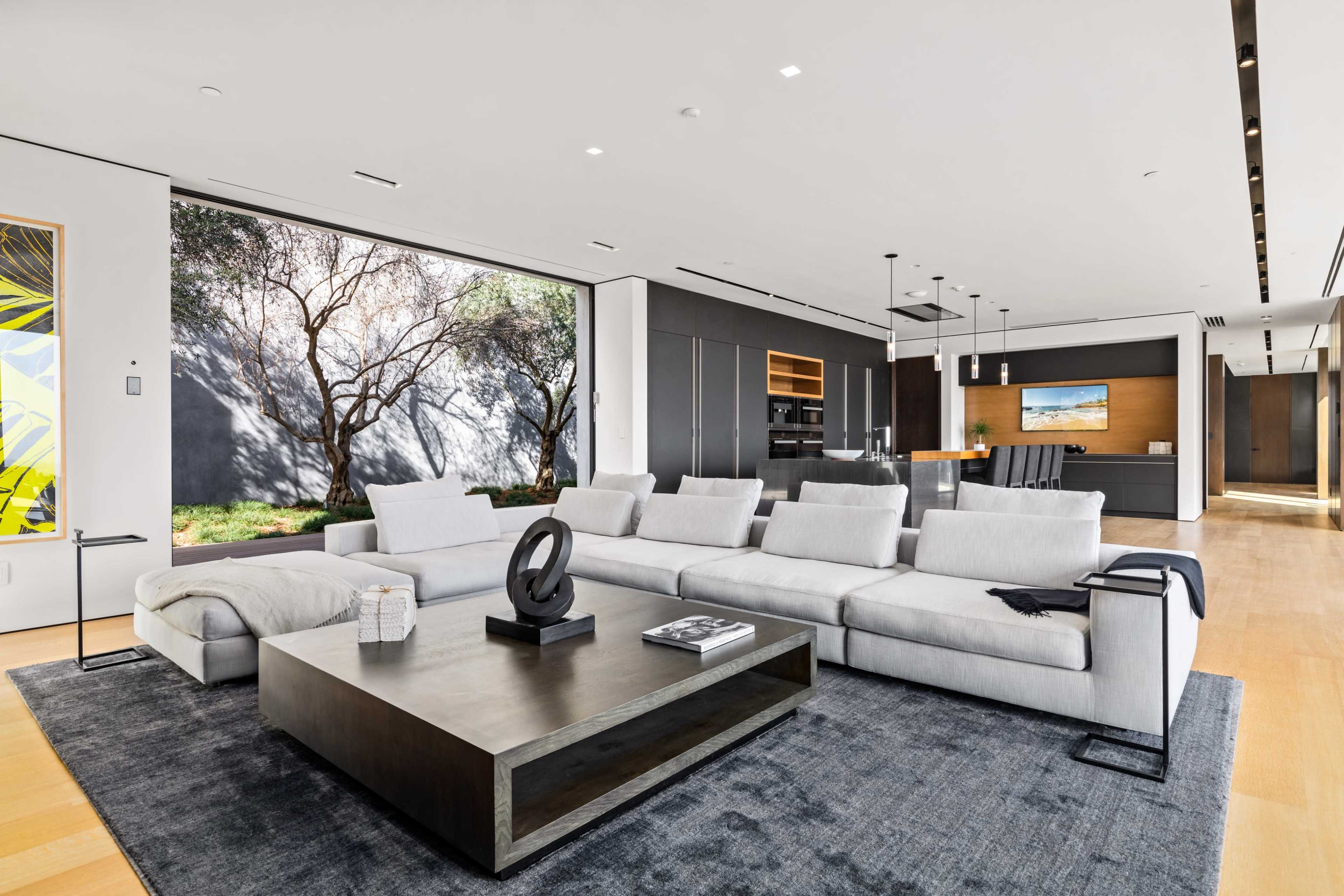 The image depicts a modern living room featuring a sleek gray sectional sofa, a dark coffee table, and large windows that allow natural light to illuminate the space.