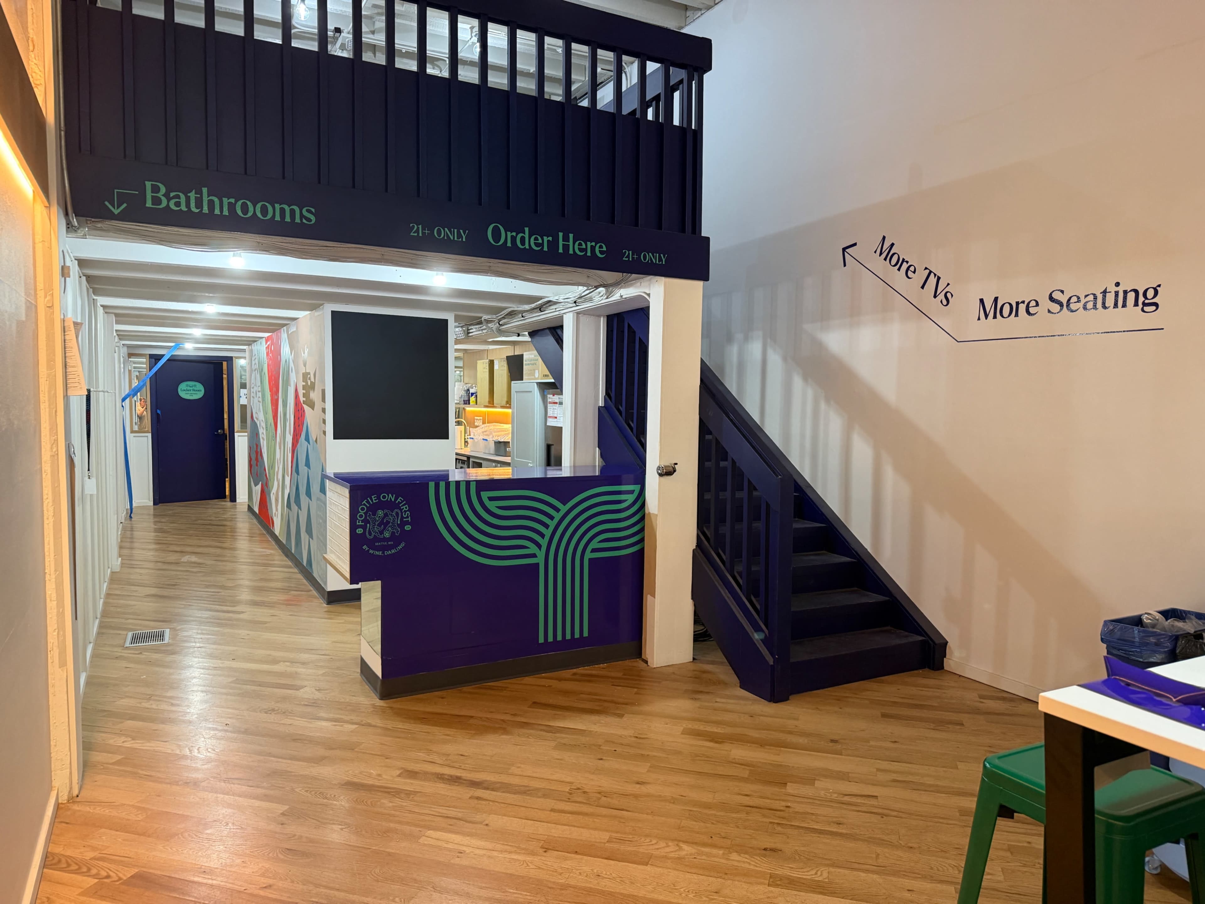 The image shows a brightly lit hallway in a building with signage directing to bathrooms and an order area, featuring wooden floors and a staircase leading up.