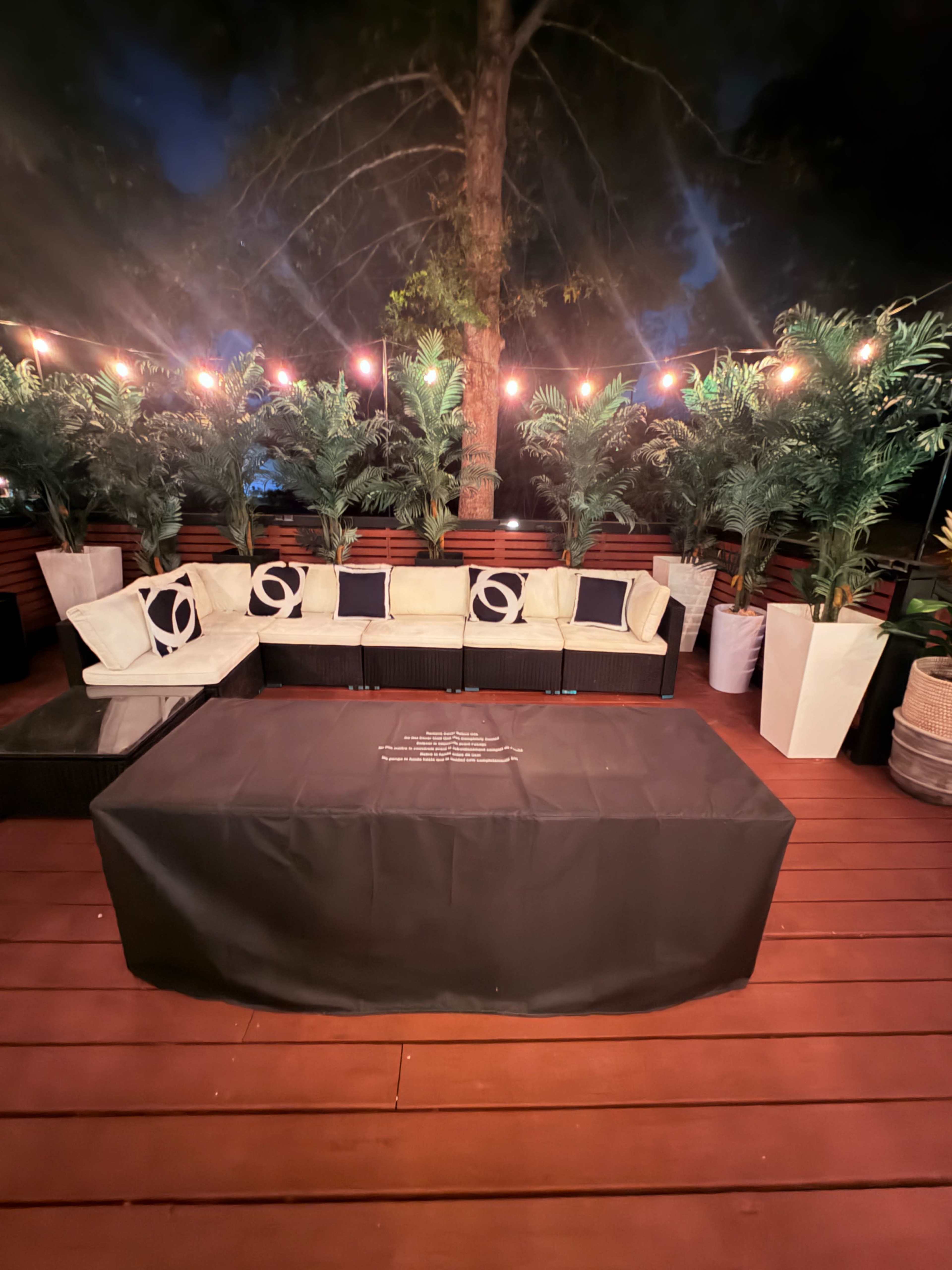 The image shows a seating area featuring a U-shaped couch with decorative cushions, surrounded by potted plants and string lights in a dimly lit outdoor space.