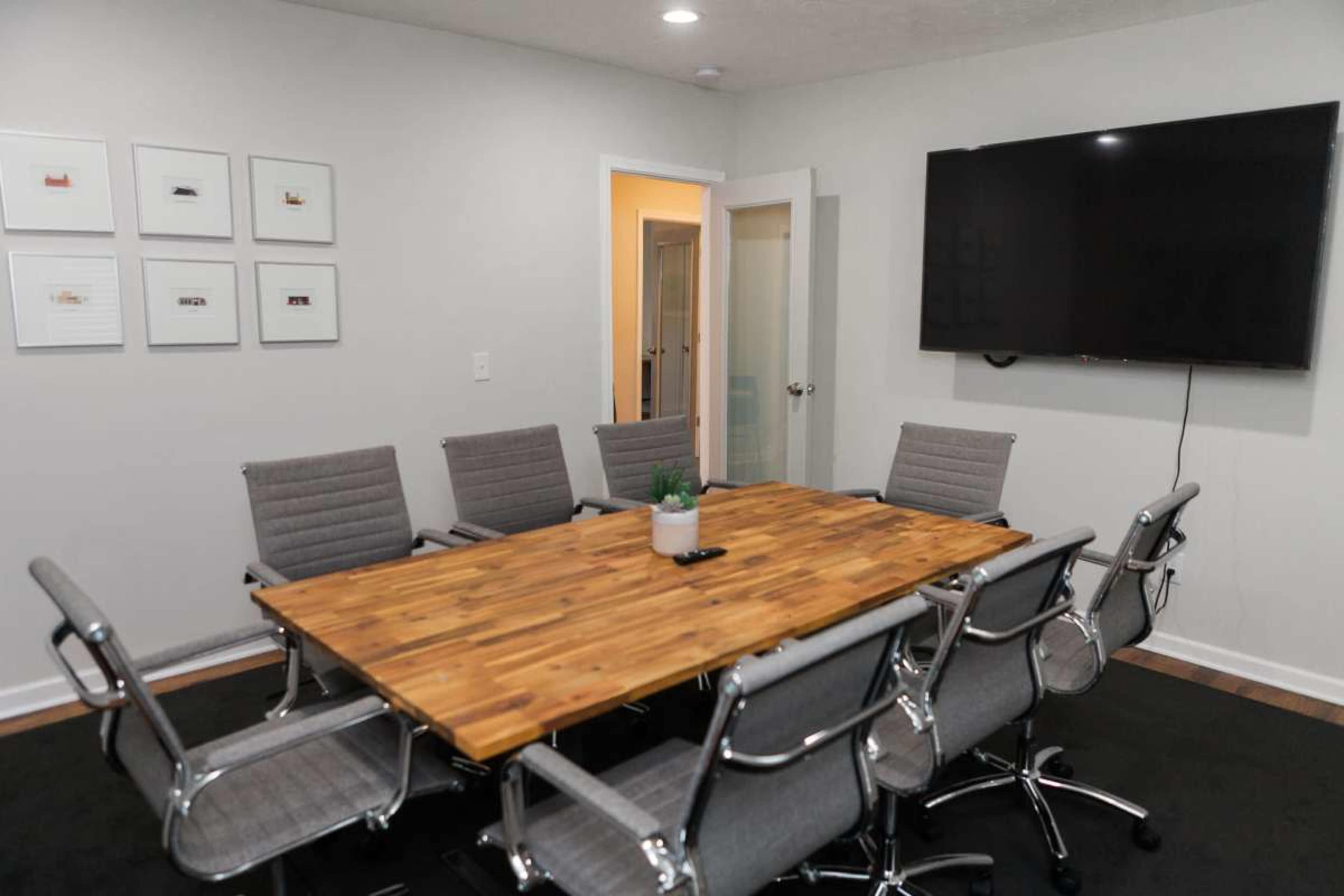 The image shows a modern conference room with a large wooden table surrounded by eight gray chairs and a wall-mounted television.