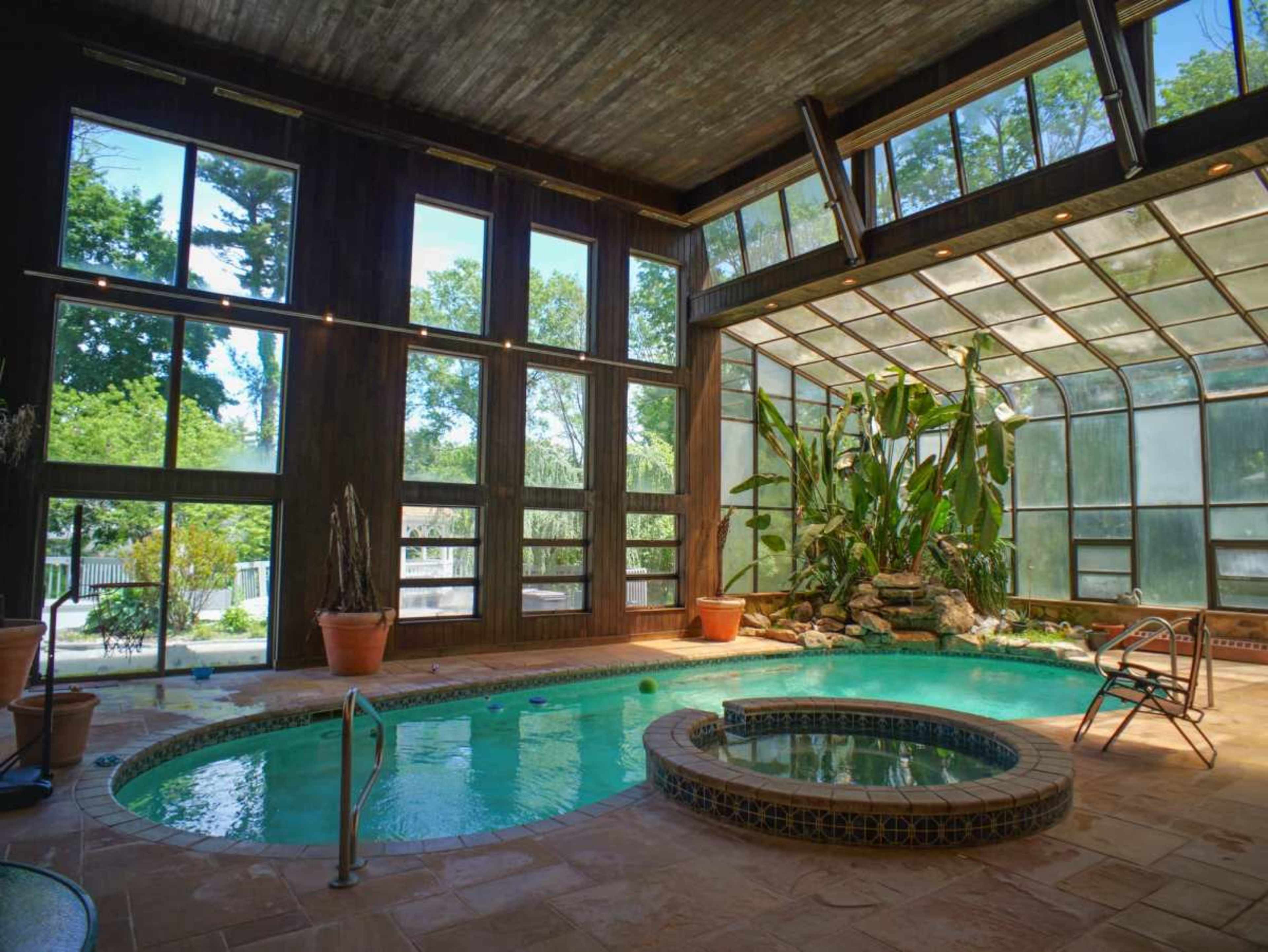 The image shows an indoor swimming pool area with large windows allowing natural light, surrounded by potted plants and a small hot tub.