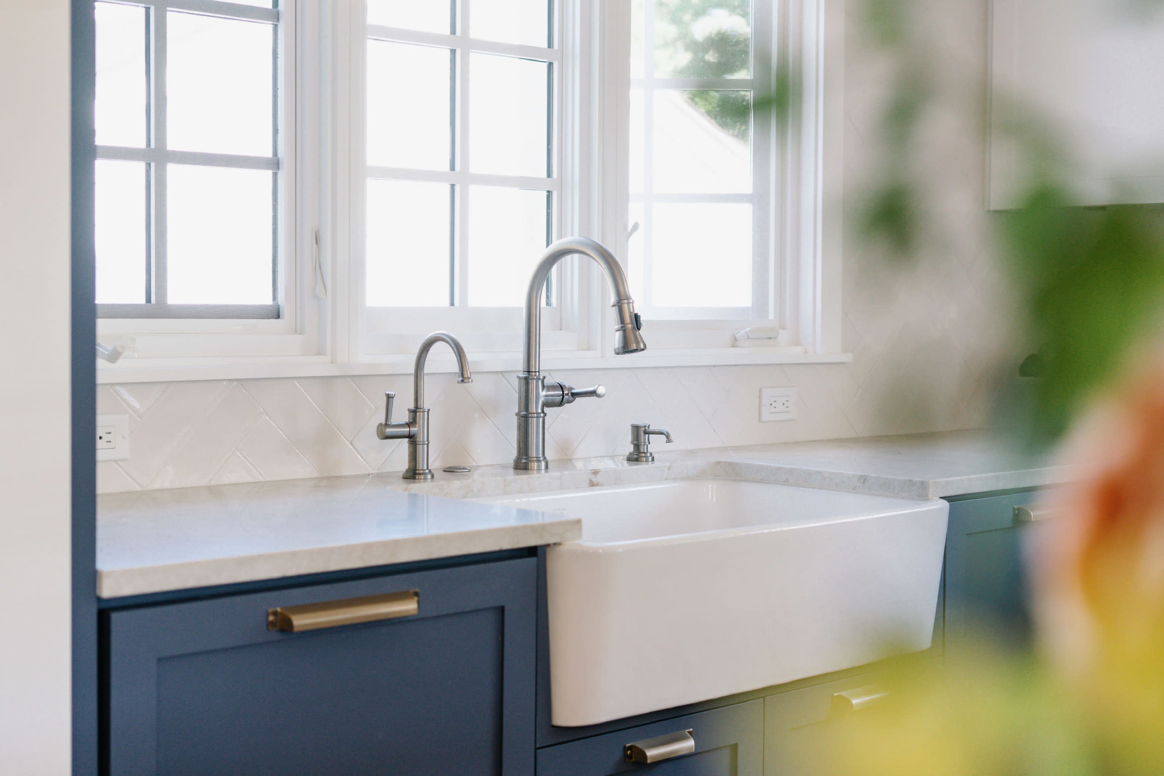 A farmhouse sink with a pull-down faucet is positioned beneath three windows, surrounded by a countertop and navy blue cabinetry.