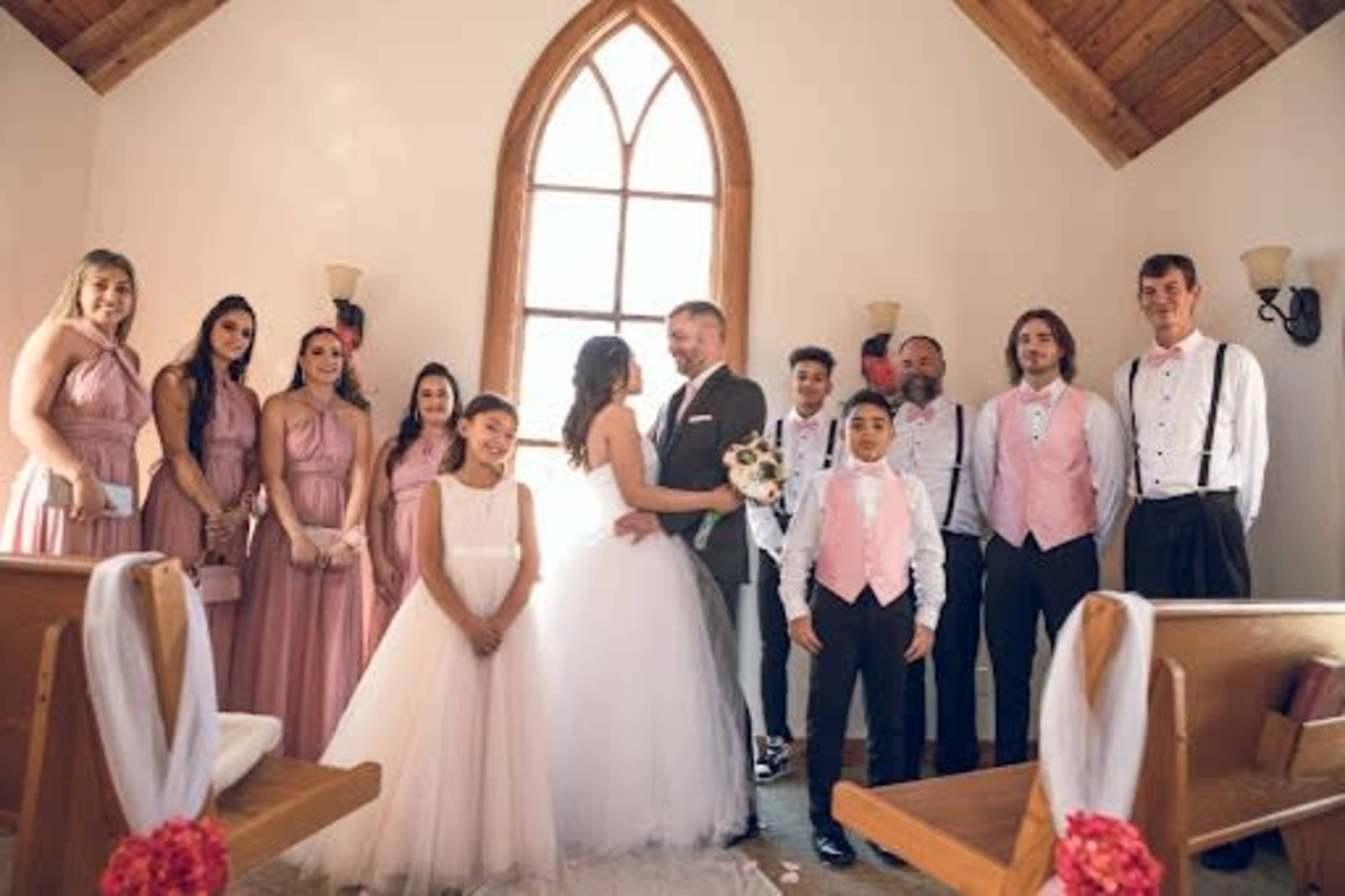 A couple stands at the altar surrounded by their wedding party, all dressed in formal attire inside a chapel with a large arched window.