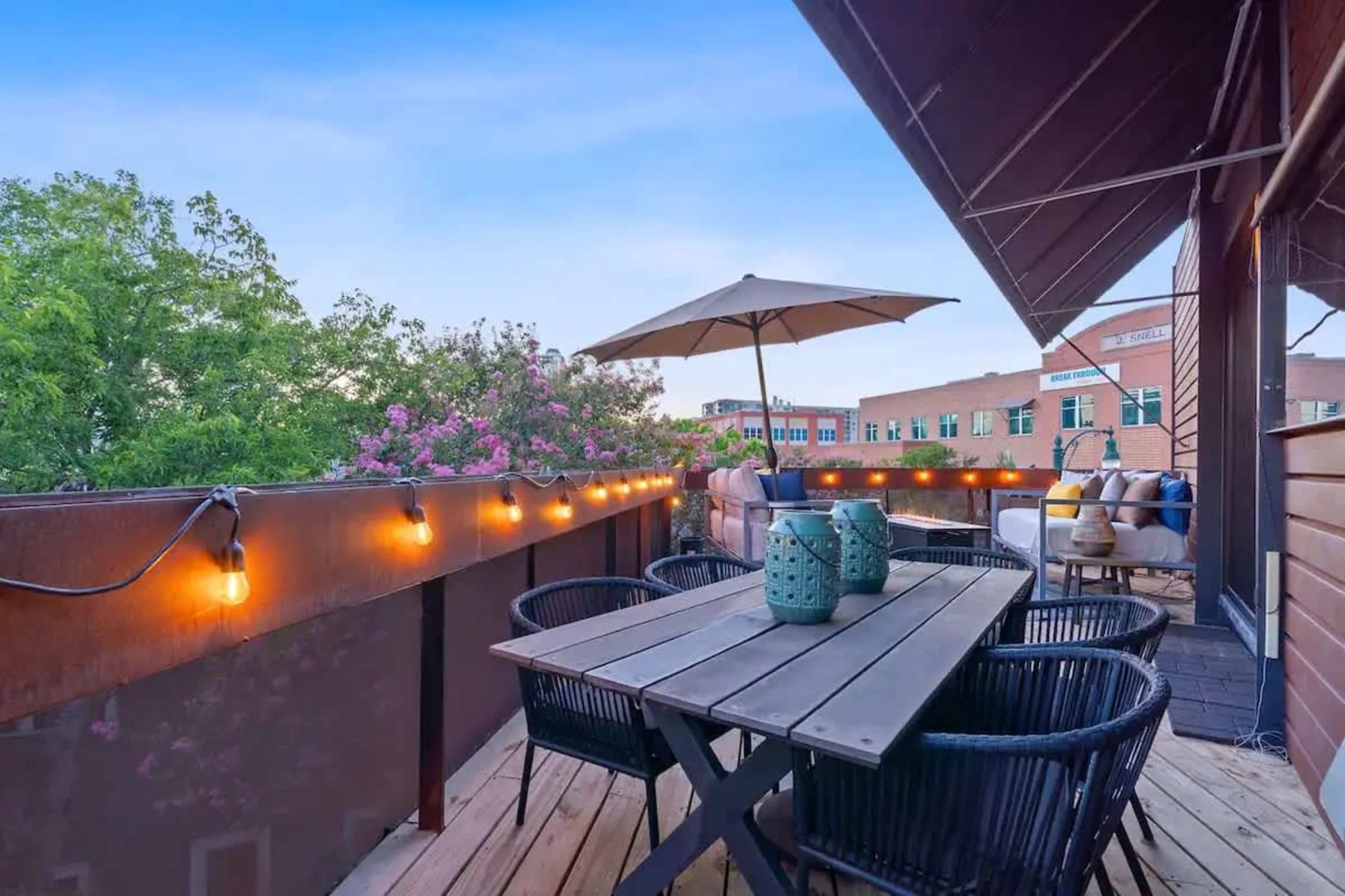A rooftop terrace features a wooden table surrounded by black wicker chairs, with string lights and an umbrella, overlooking greenery and a brick building.
