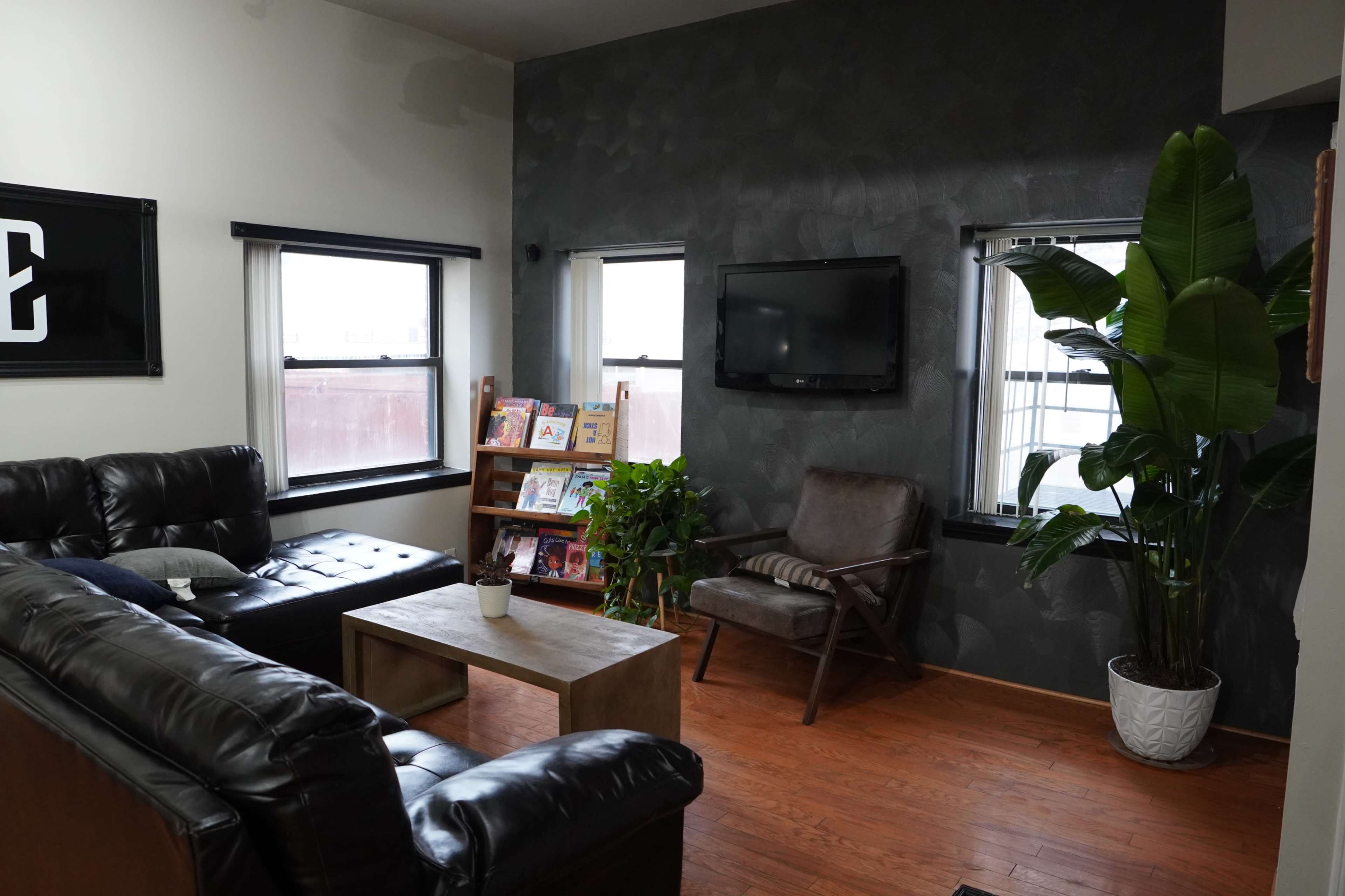 A cozy living room with a black leather couch, a wooden coffee table, a plant next to an armchair, and a wall-mounted television.
