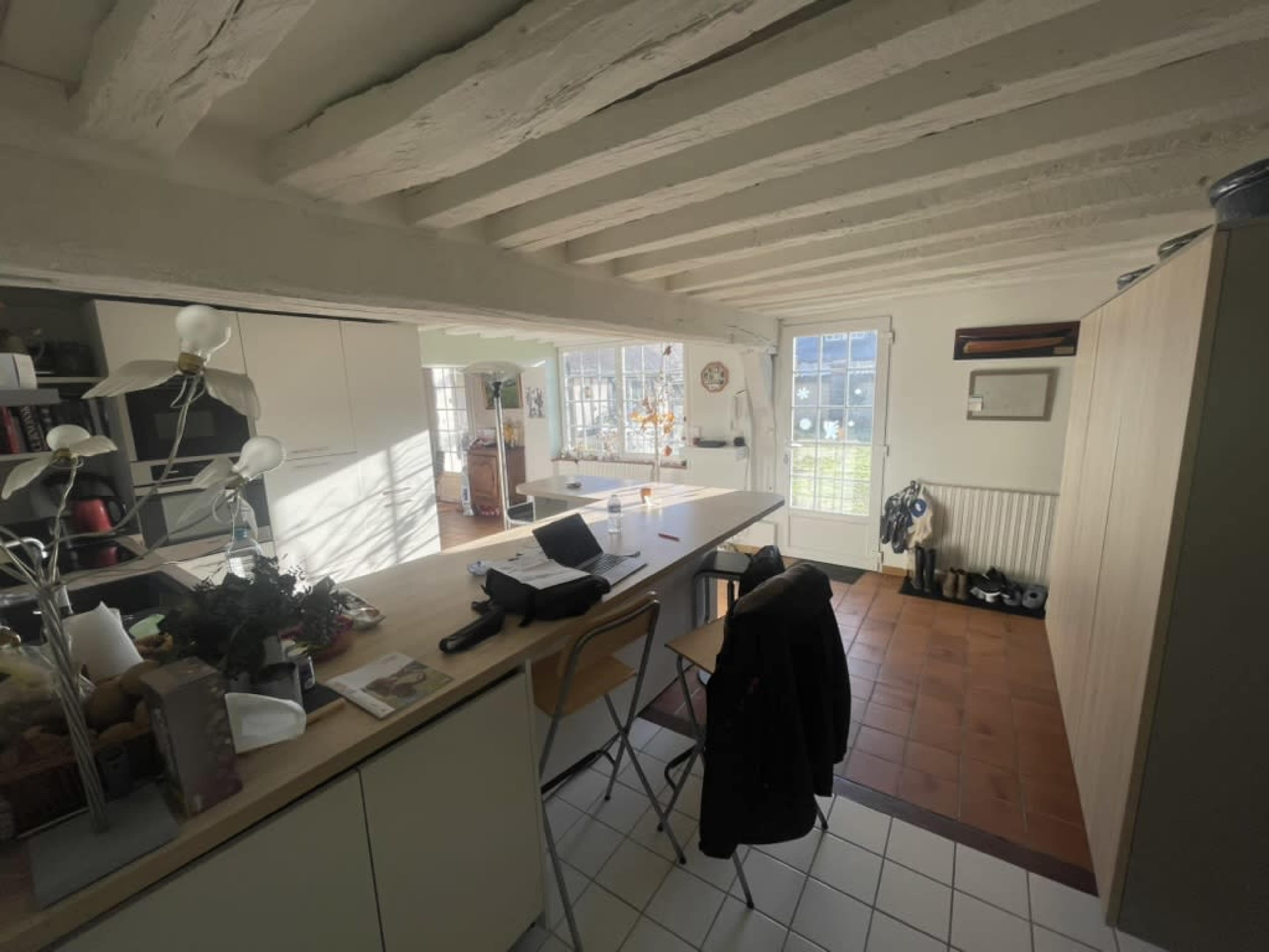 The image shows a well-lit kitchen with wooden beams, a central island, and a small dining area with chairs.