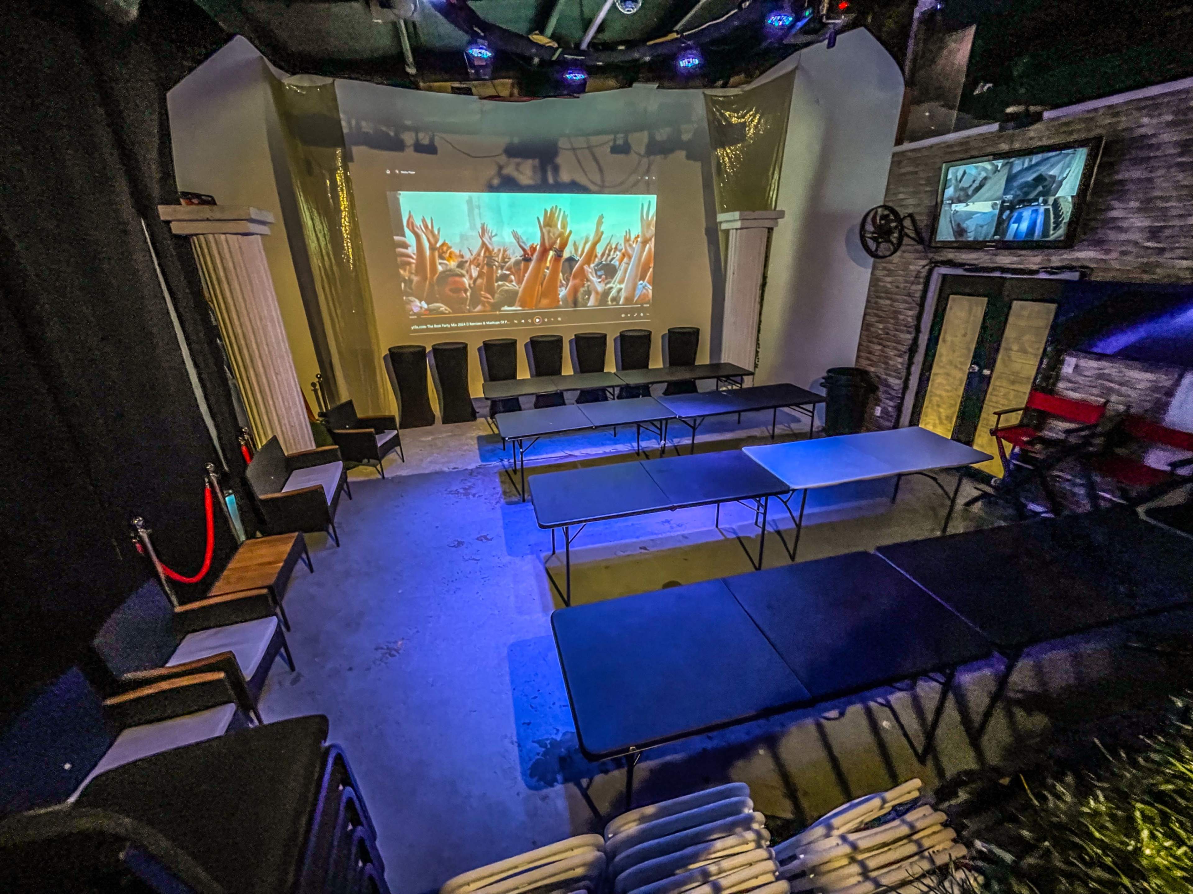 The image shows a room set up with several long tables and chairs arranged for an event, featuring a large screen displaying a vibrant audience.