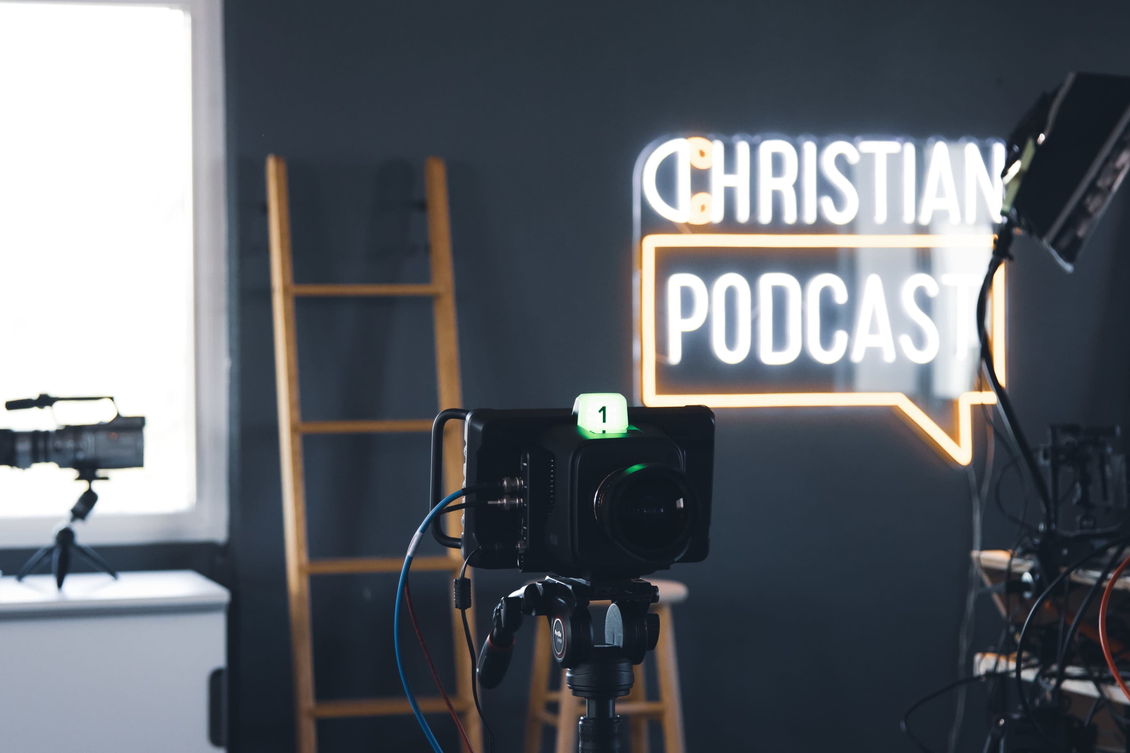A camera is set up in front of a neon sign reading "CHRISTIAN PODCAST" on a dark wall.