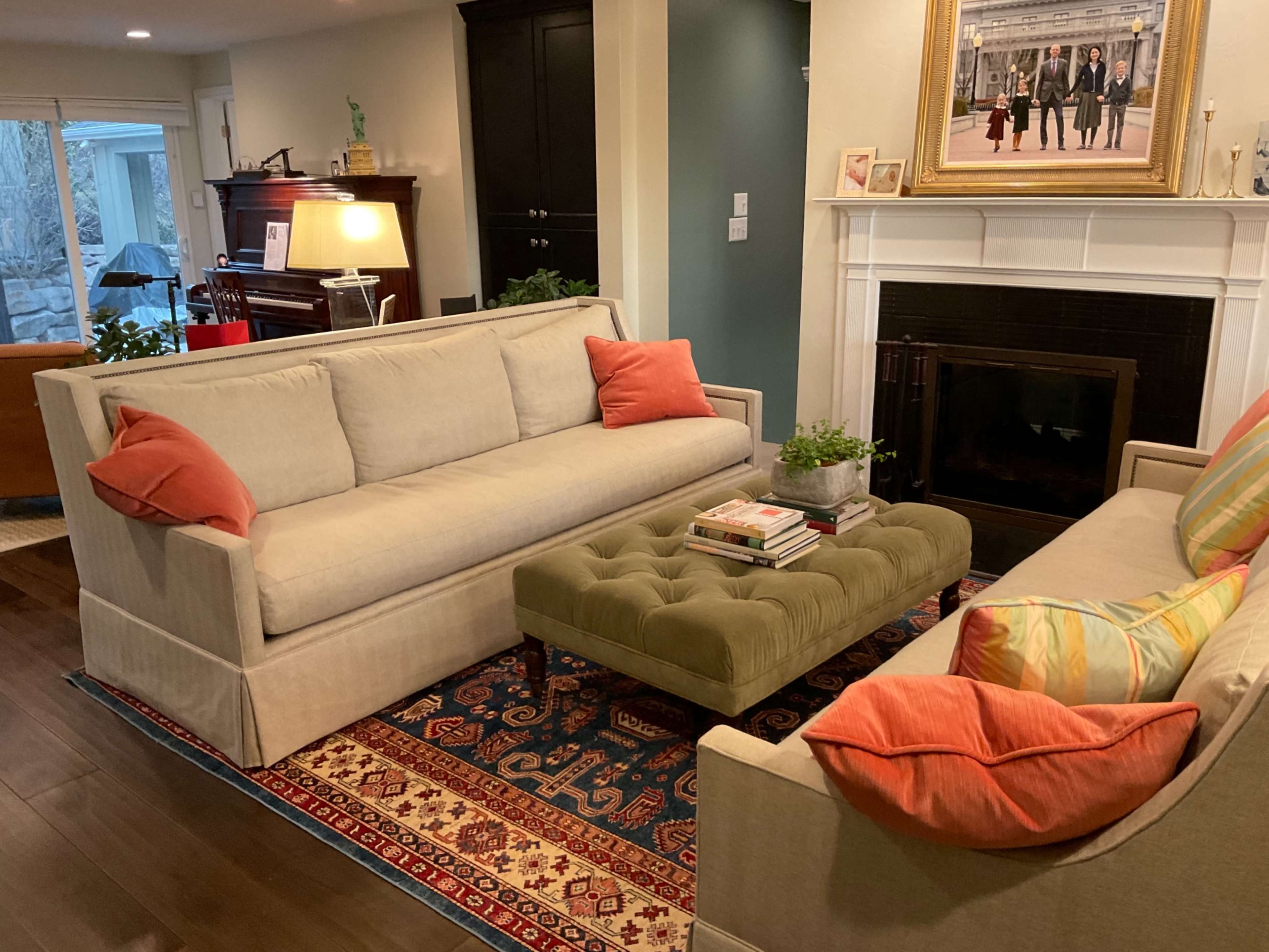 A living room with two sofas arranged around a green upholstered coffee table on a patterned rug, with a fireplace and piano in the background.