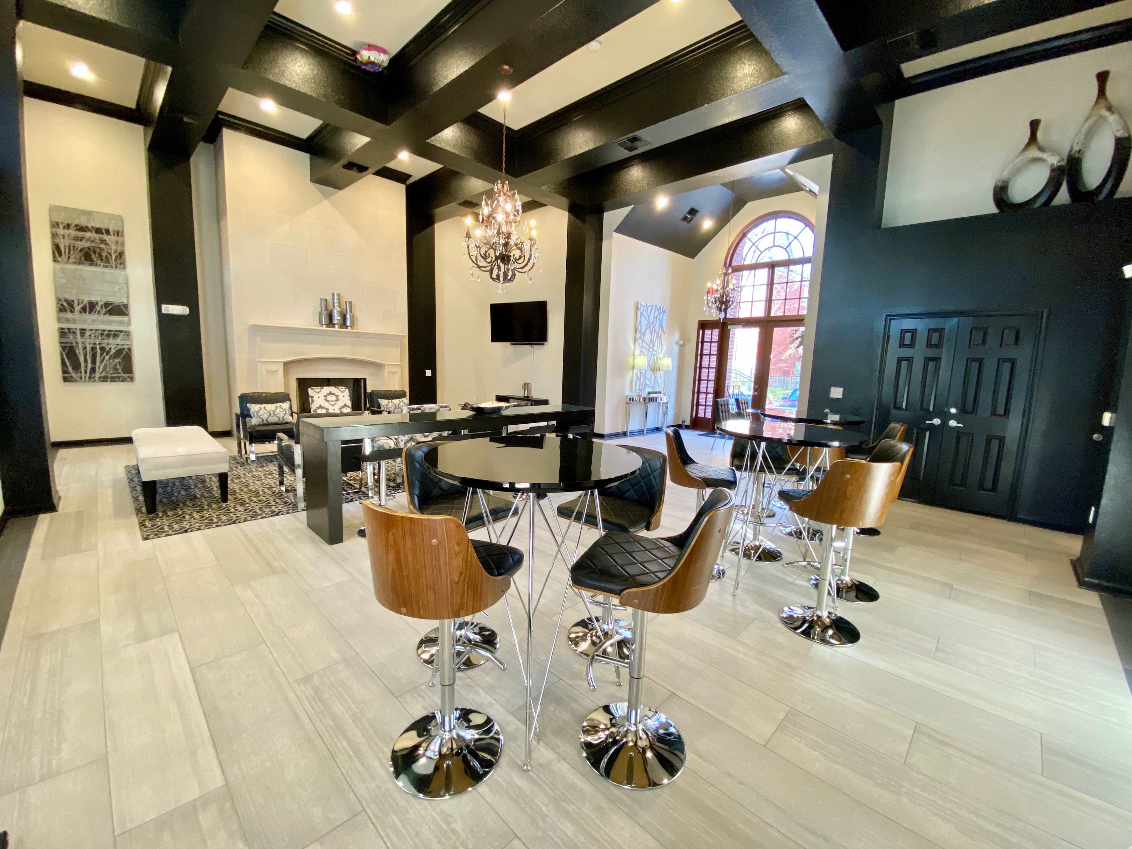 The image shows a modern indoor lounge area with a black and white color scheme, featuring a central black table surrounded by high stools, a fireplace, and large windows.