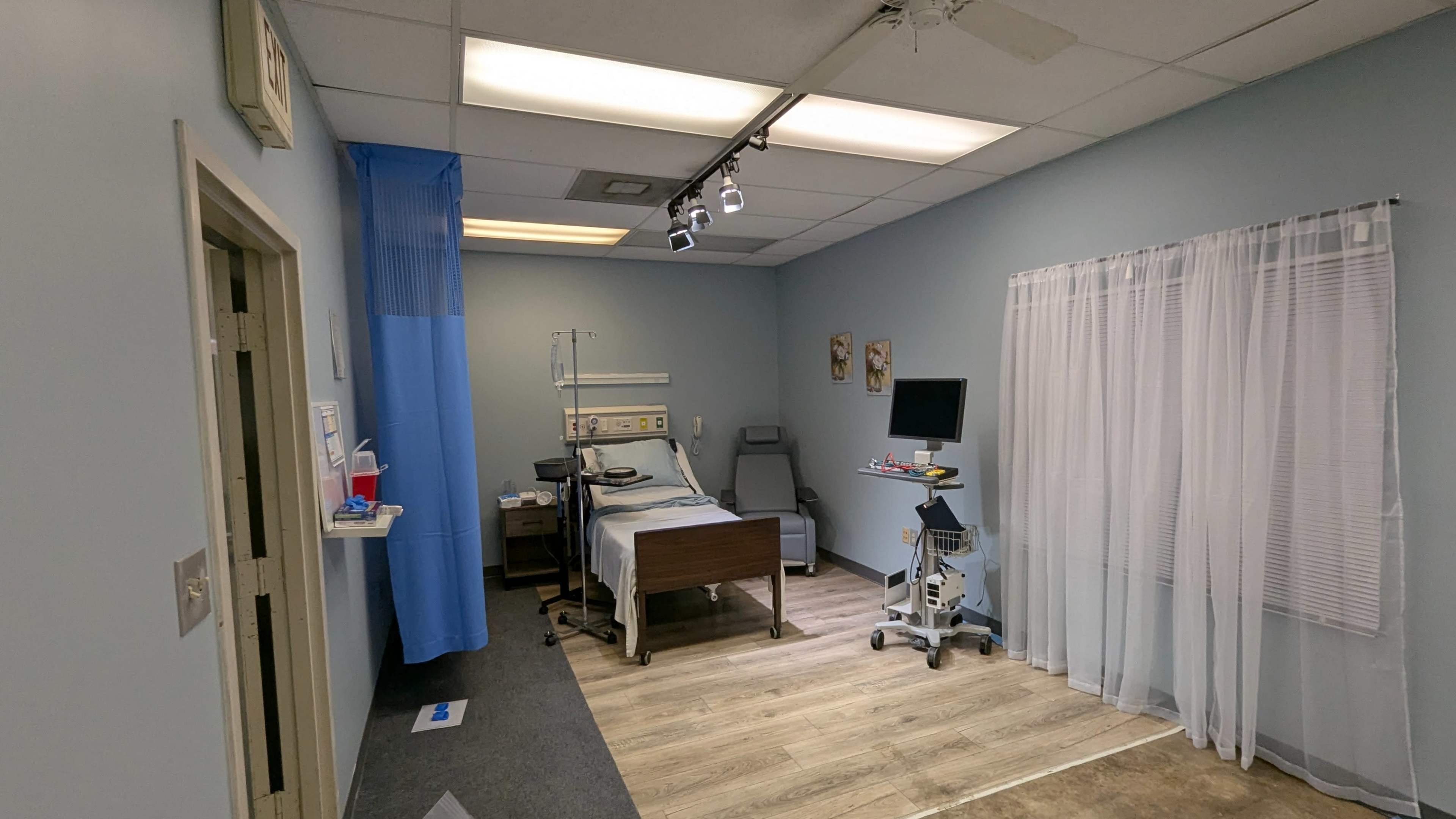 The image shows a hospital room featuring a bed, medical equipment, and a curtain, with blue walls and a wooden floor.