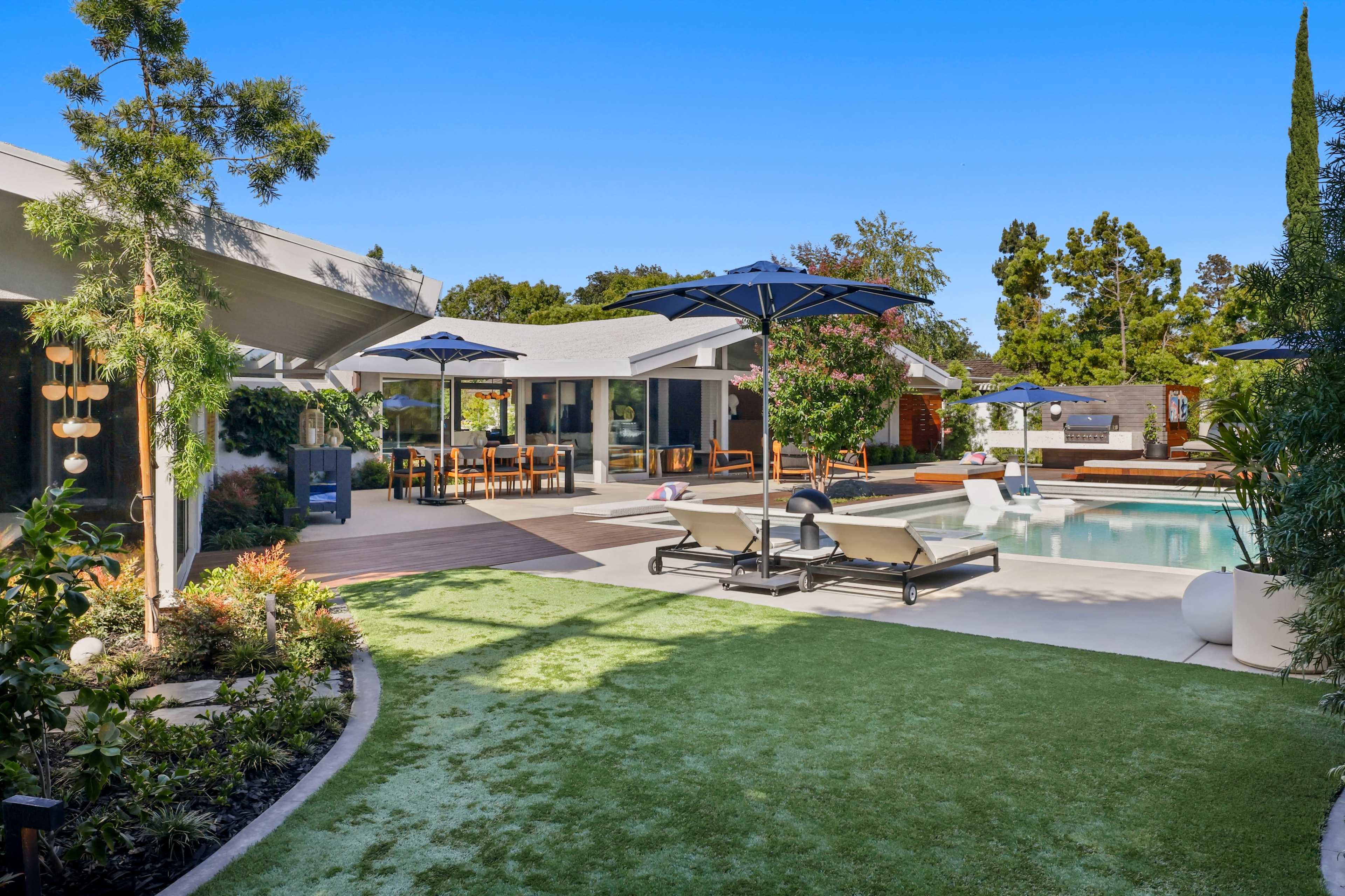 A modern backyard with a swimming pool, lounge chairs, and dining areas surrounded by greenery and patio umbrellas.