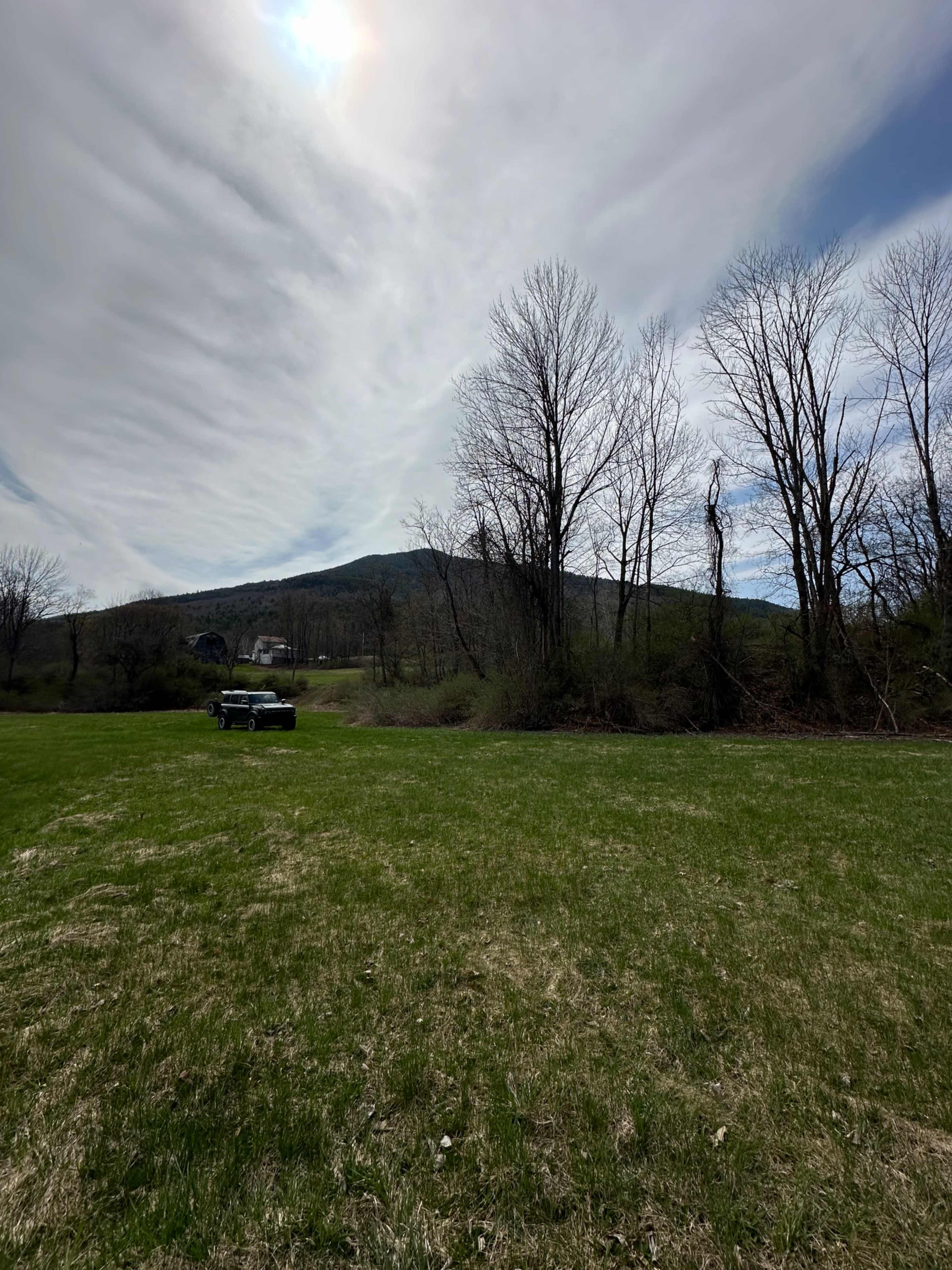A black vehicle is parked on a grassy field with bare trees and a hill in the background under a cloudy sky.