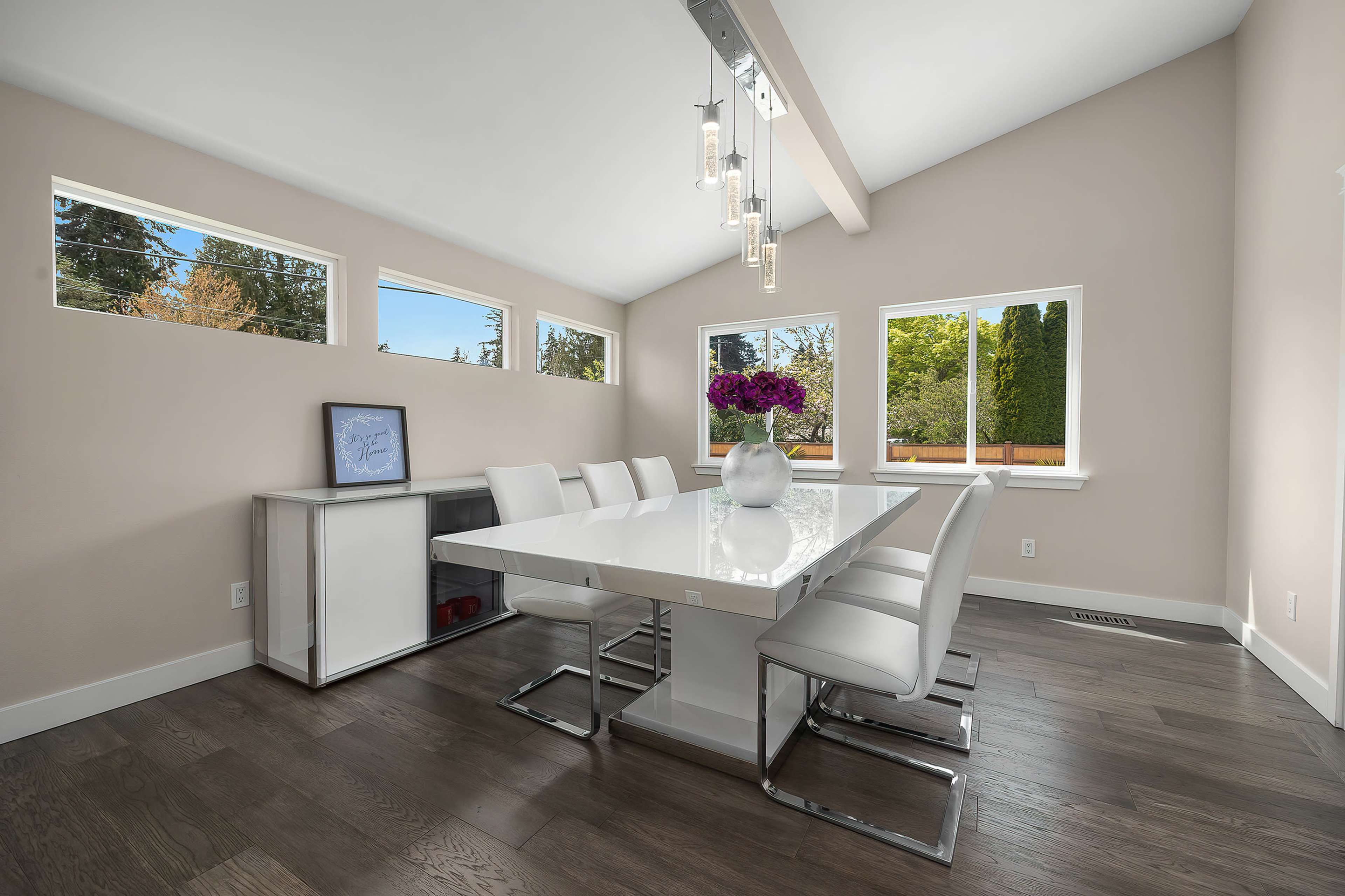 A modern dining room features a white table surrounded by sleek chairs, illuminated by pendant lights and complemented by large windows.