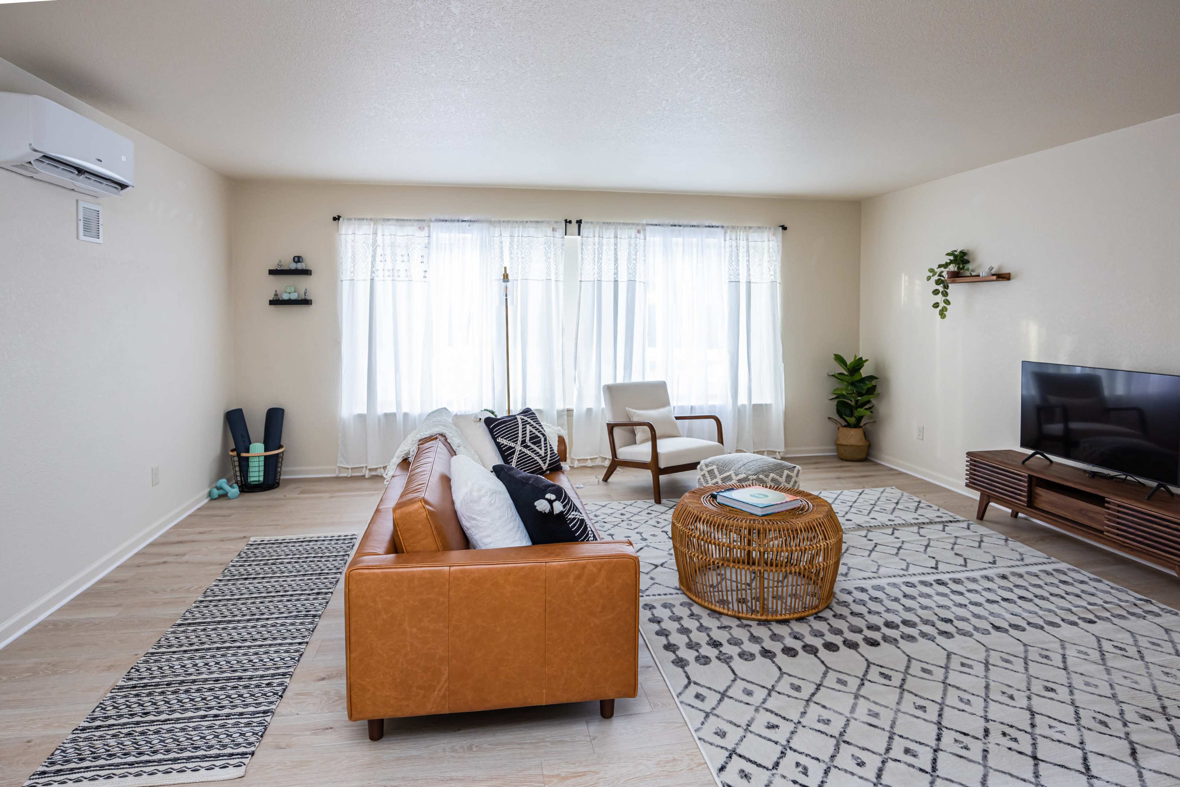 A modern living room features a brown leather sofa, a woven coffee table, and light-colored walls, with large windows draped in white curtains allowing natural light.