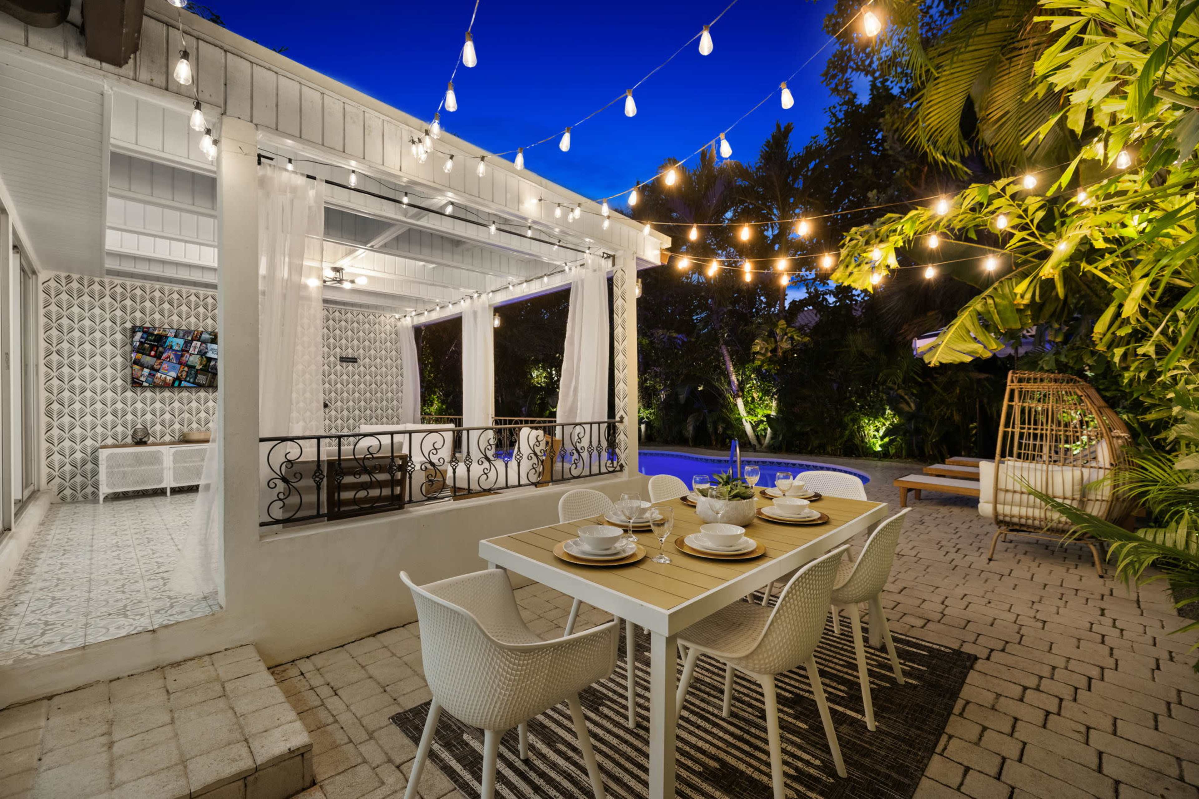 A modern patio features a dining table set with tableware under string lights, surrounded by lush greenery and a swimming pool in the background.